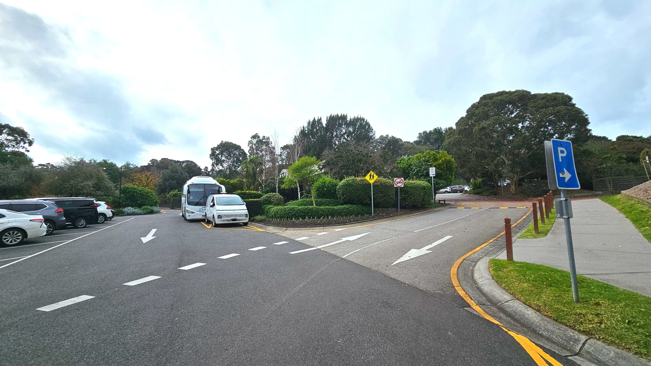 A car park area with several vehicles parked on the left side, including a large white bus and a white van. The road is marked with white arrows indicating traffic direction. A blue parking sign with a right arrow is visible on the right, next to a paved footpath bordered by wooden posts and grass. Dense greenery and trees form a natural boundary at the back, with pedestrian and no entry signs visible.