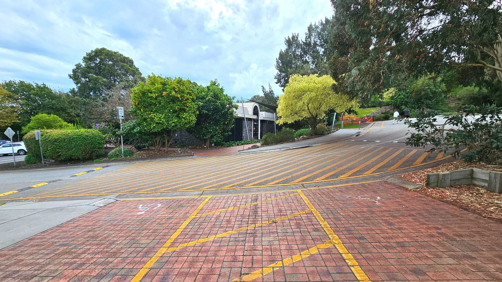 A sloped road with yellow diagonal lines leads up to a building partially obscured by lush greenery. The road is bordered by a brick-paved area with yellow markings in the foreground. To the right, a path curves upwards, flanked by trees and bushes. A signpost with 'END' is visible near the centre. The area feels open with scattered trees and a few parked cars visible on the left.