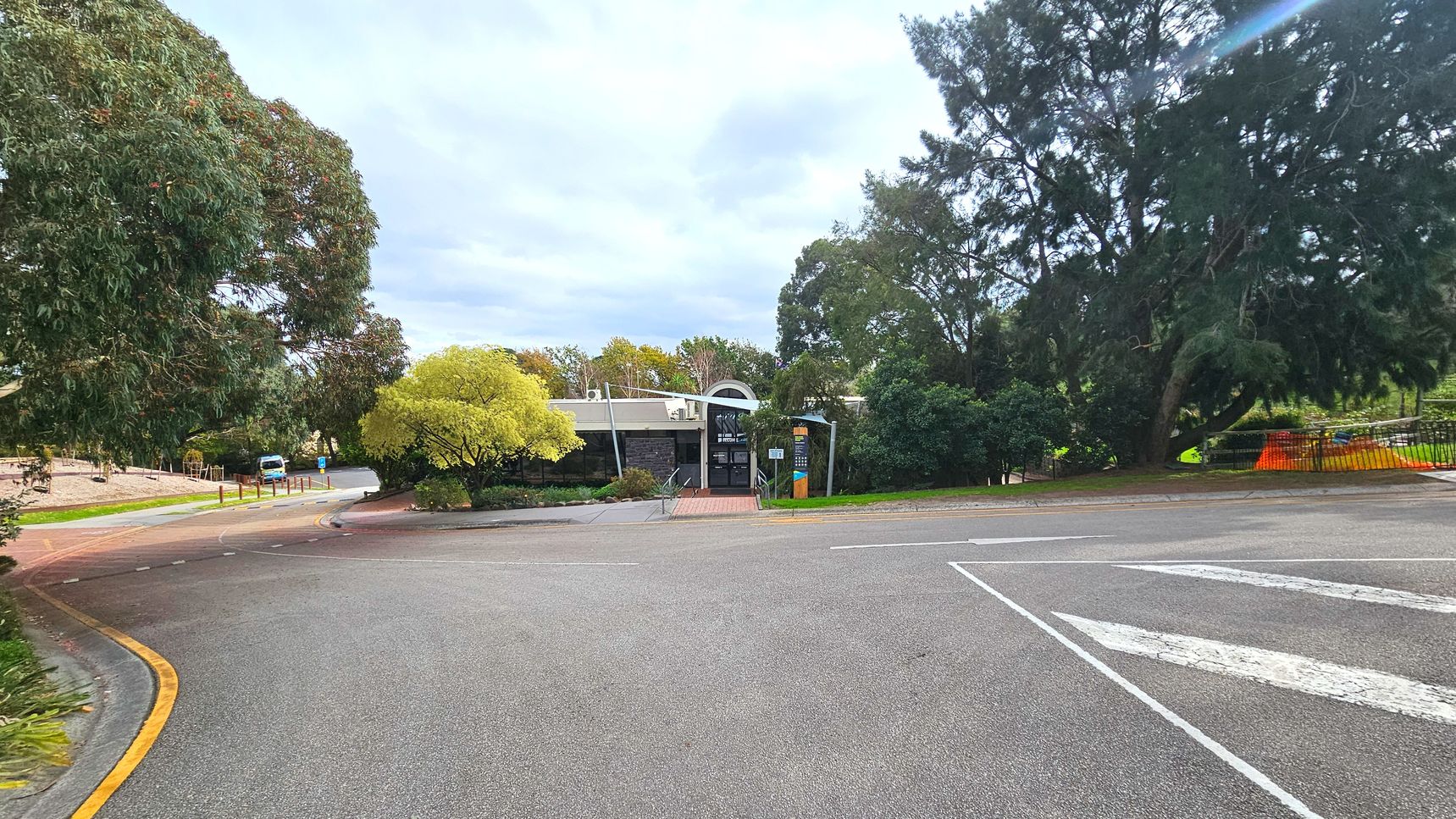 A single-storey building with a flat roof and a central arched entrance is surrounded by lush greenery. A paved path leads to the entrance, flanked by a tall signpost. The foreground features a wide asphalt road with a yellow kerb line and white directional arrows. Large trees frame the scene on both sides, creating a semi-enclosed feel. A parked vehicle is visible to the left near a series of bollards.