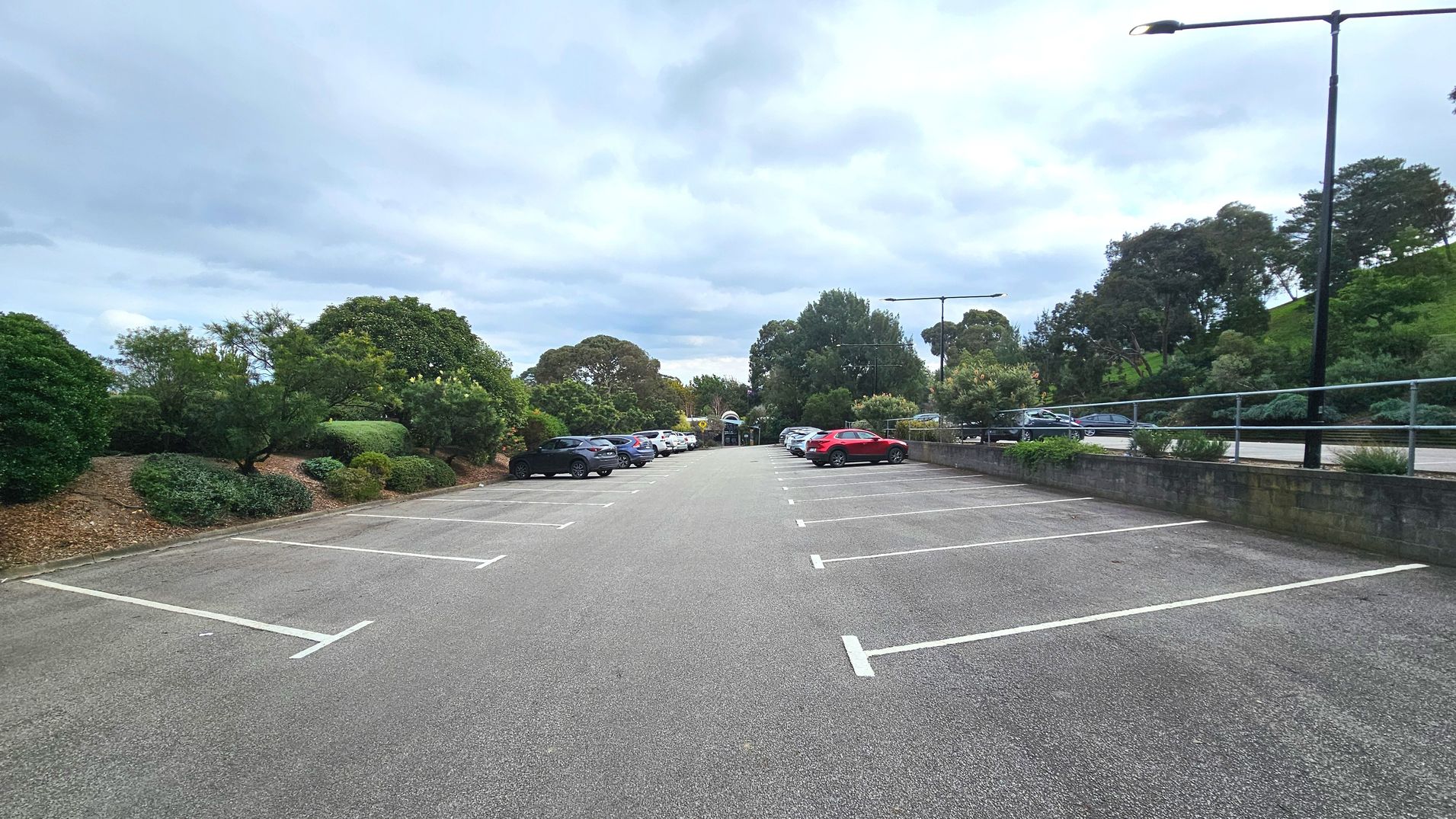 A spacious car park with marked parking bays on an asphalt surface. Several cars are parked on the left side, while the right side has fewer vehicles. The area is bordered by lush greenery, including trees and shrubs, providing a natural boundary. Tall street lamps line the right side, and a low brick wall with a metal railing runs parallel to the parking area, enhancing the sense of enclosure.