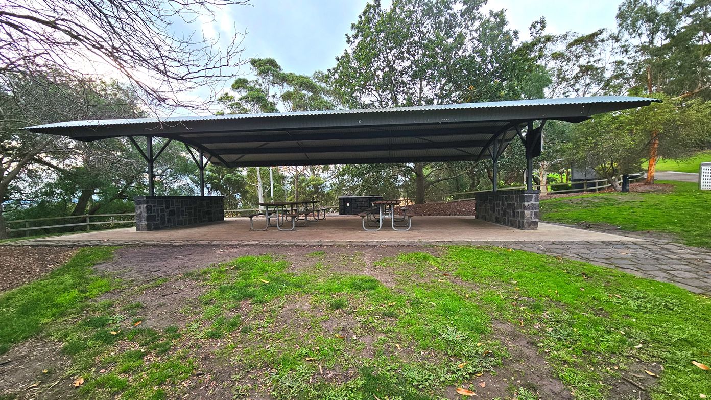 A covered picnic area with a corrugated metal roof supported by black metal beams and stone pillars. Beneath the shelter are several picnic tables with metal frames and wooden tops. The ground is a mix of grass and dirt, transitioning to a paved stone path leading to the shelter. Surrounding the area are trees and a wooden fence, creating a semi-enclosed space. A bin is visible to the right.