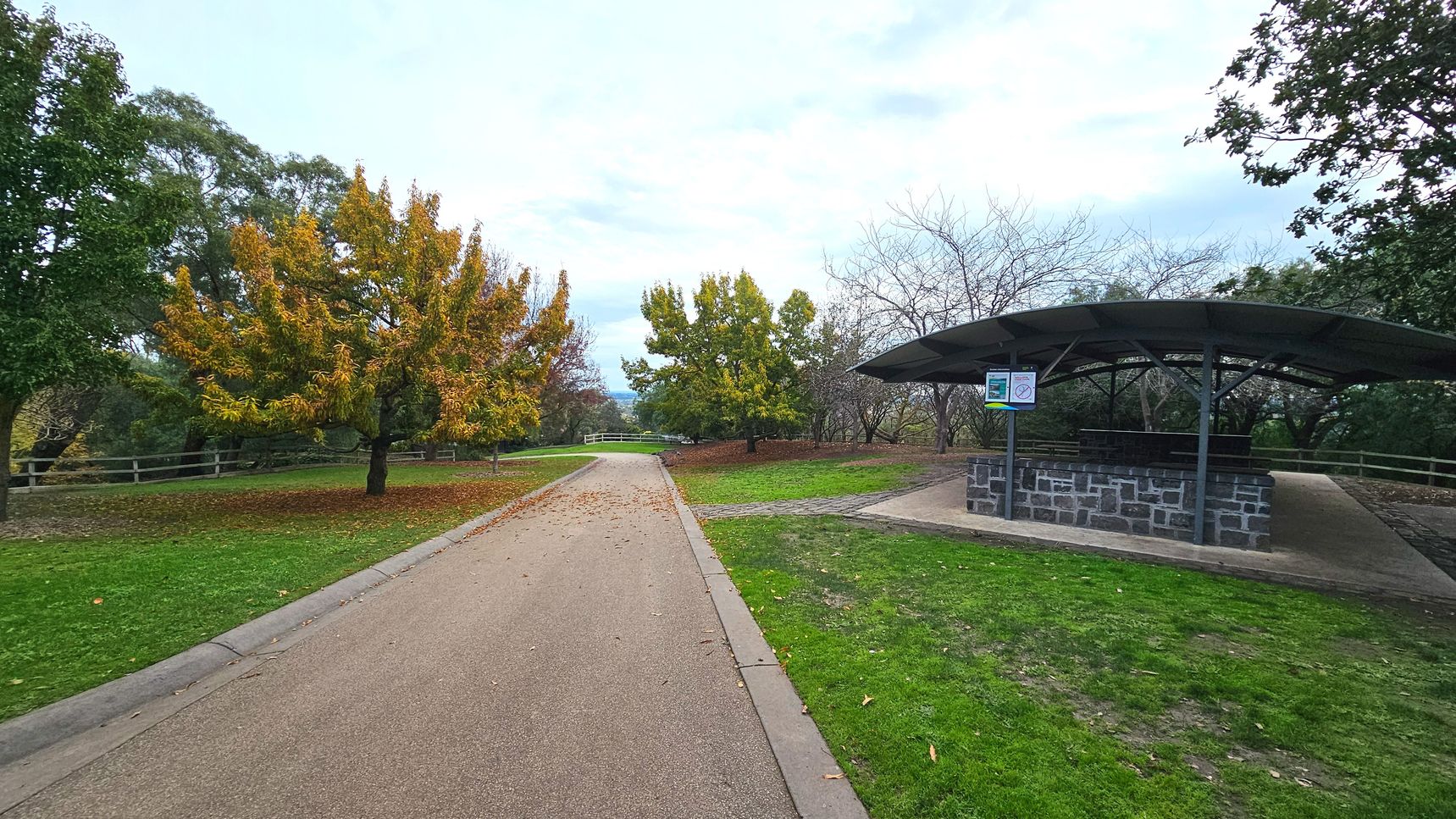 A park scene with a wide, paved path running through the centre, flanked by green grass and autumnal trees with yellow and orange leaves. To the right, a stone and metal shelter with a curved roof provides seating, indicated by a sign. A wooden fence lines the background, enclosing the area. The ground transitions from grass to pavement smoothly, and the space feels open with trees scattered throughout.