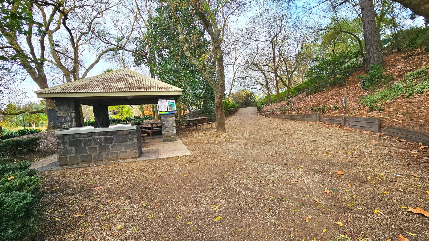 A park scene with a stone pavilion on the left, featuring a corrugated metal roof supported by stone pillars. Inside, picnic tables are visible. A dirt path leads away from the pavilion, flanked by trees and a low wooden barrier on the right. The ground is covered with fallen leaves, and the area is surrounded by dense foliage, creating a semi-enclosed feel. A sign is posted on the pavilion wall.