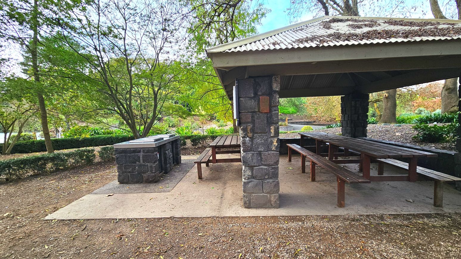 A sheltered picnic area with a corrugated metal roof supported by stone pillars. Under the shelter are wooden picnic tables with benches. Adjacent to the shelter is a stone barbecue grill. The ground is a mix of concrete and dirt, with fallen leaves scattered around. Surrounding the area are lush green trees and shrubs, creating a natural boundary.