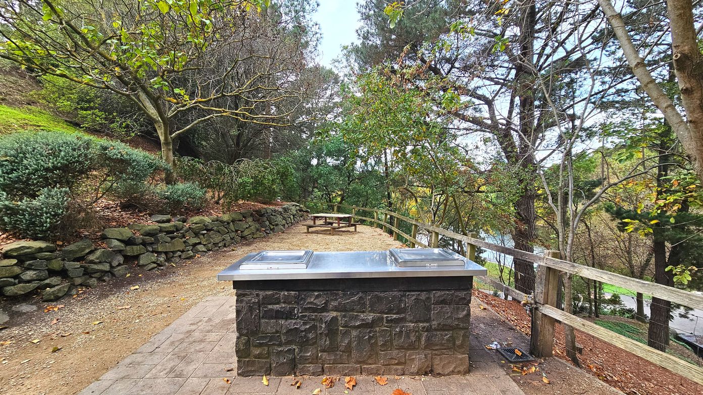 A stone barbecue with a stainless steel top is centrally placed on a paved area, surrounded by a natural setting. To the left, a low stone wall borders a dirt path lined with bushes and trees. On the right, a wooden fence runs parallel to the path, overlooking a slope with more trees. In the background, a wooden picnic table is visible, partially shaded by the surrounding foliage.