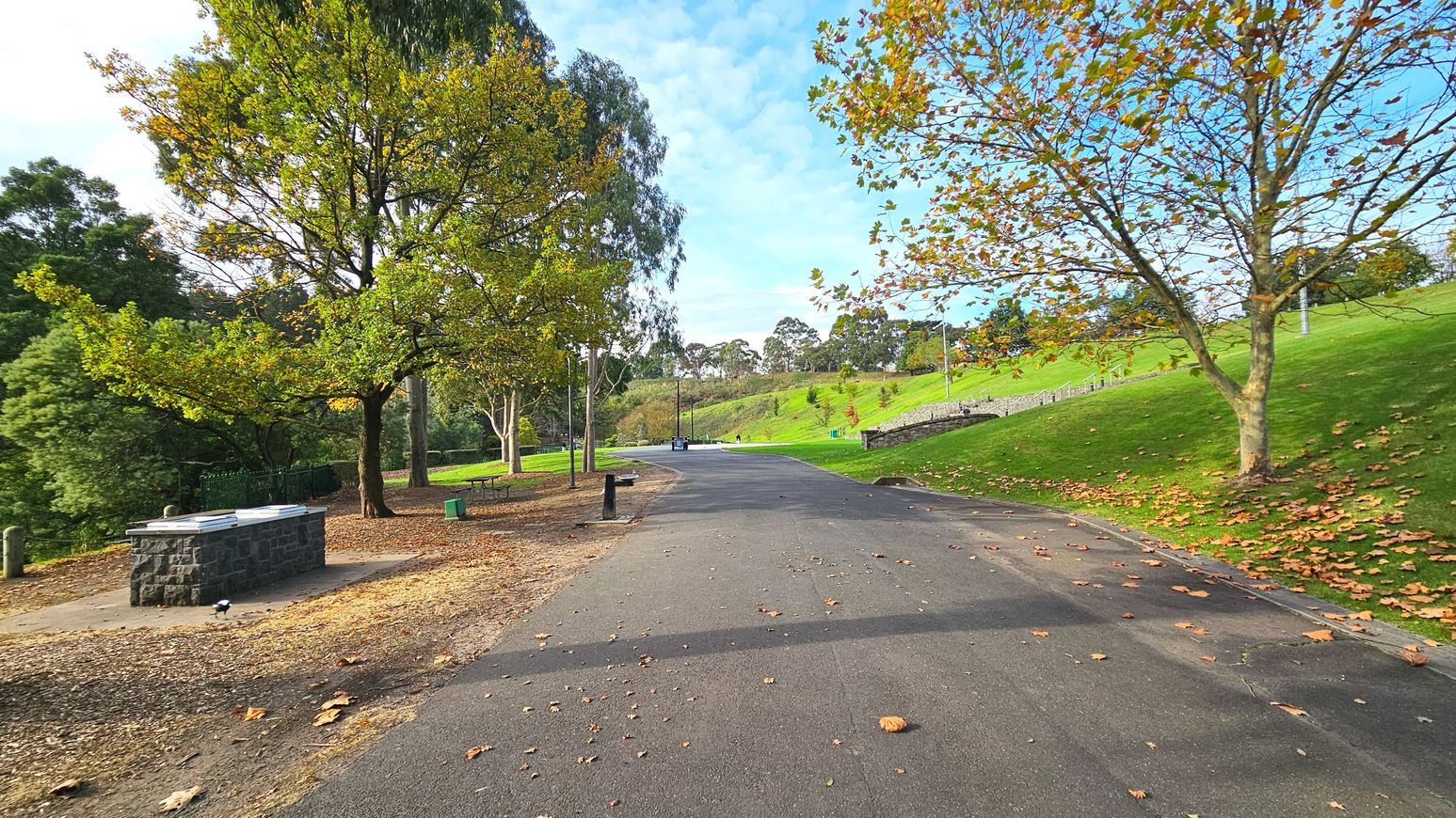 A paved path winds through a park with lush green grass on either side. Mature trees with autumn leaves line the path, providing shade. On the left, a stone barbecue area is visible, along with a picnic table. The path is clear and wide, with a gentle slope upwards. A few benches and bins are scattered along the route, and a low stone wall runs parallel to the path on the right.