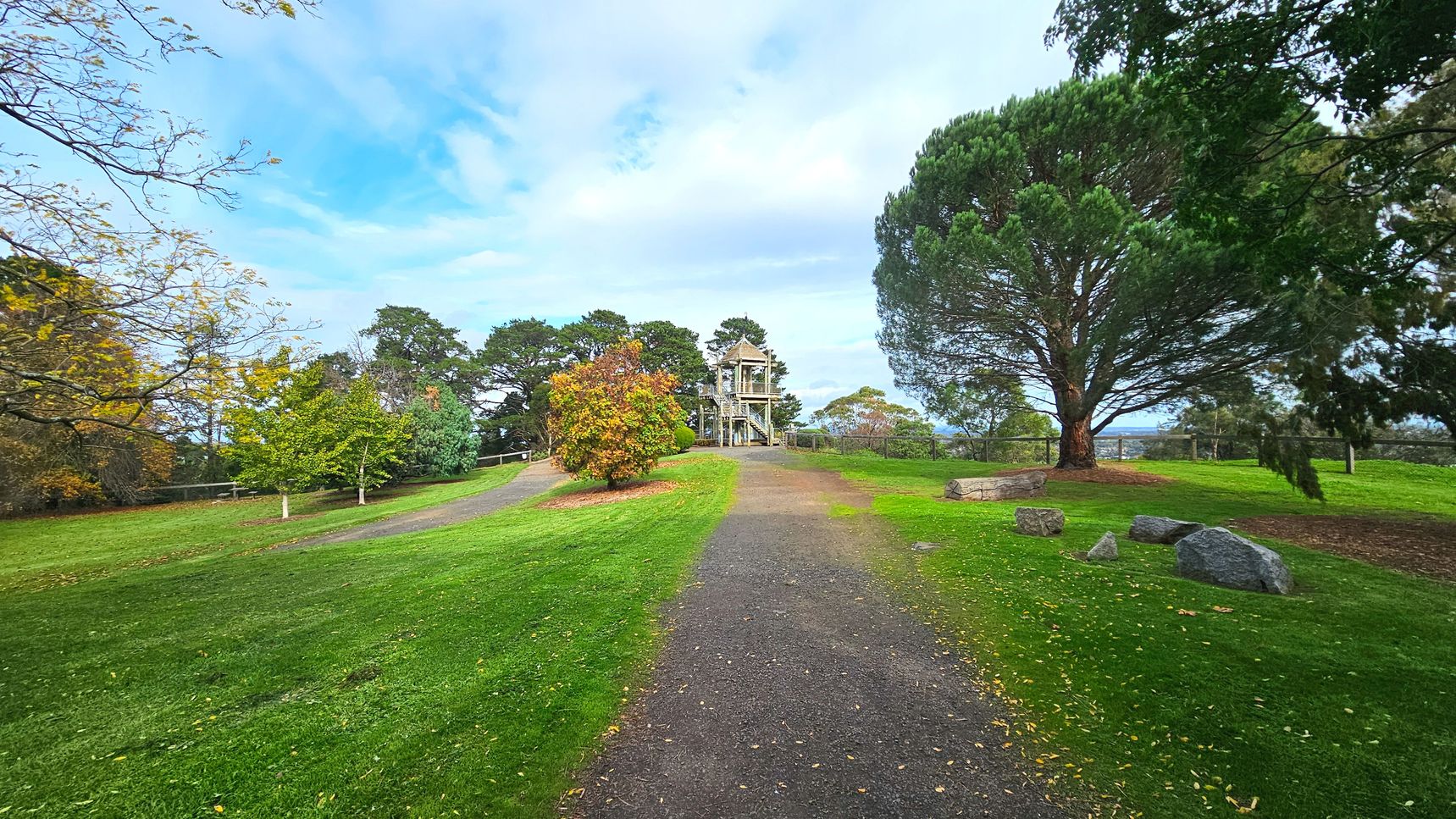 A park scene with a gravel path leading towards a wooden observation tower in the centre, surrounded by lush green grass and various trees. The tower has a pointed roof and is flanked by a mix of deciduous and evergreen trees. Large rocks and a log are scattered on the grass to the right. The area is open with a few trees providing partial enclosure, and a wooden fence is visible in the background.