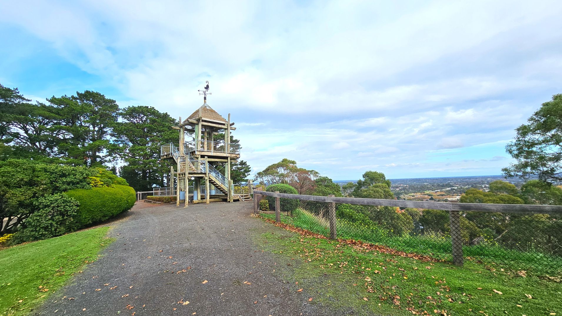 A wooden observation tower with a pointed roof stands prominently on a grassy hilltop. It features a spiral staircase leading to an upper platform. To the left, dense green bushes and trees provide a natural backdrop. A gravel path runs alongside the tower, bordered by a wooden and wire mesh fence overlooking a distant townscape. Fallen leaves are scattered on the ground, indicating a seasonal transition.