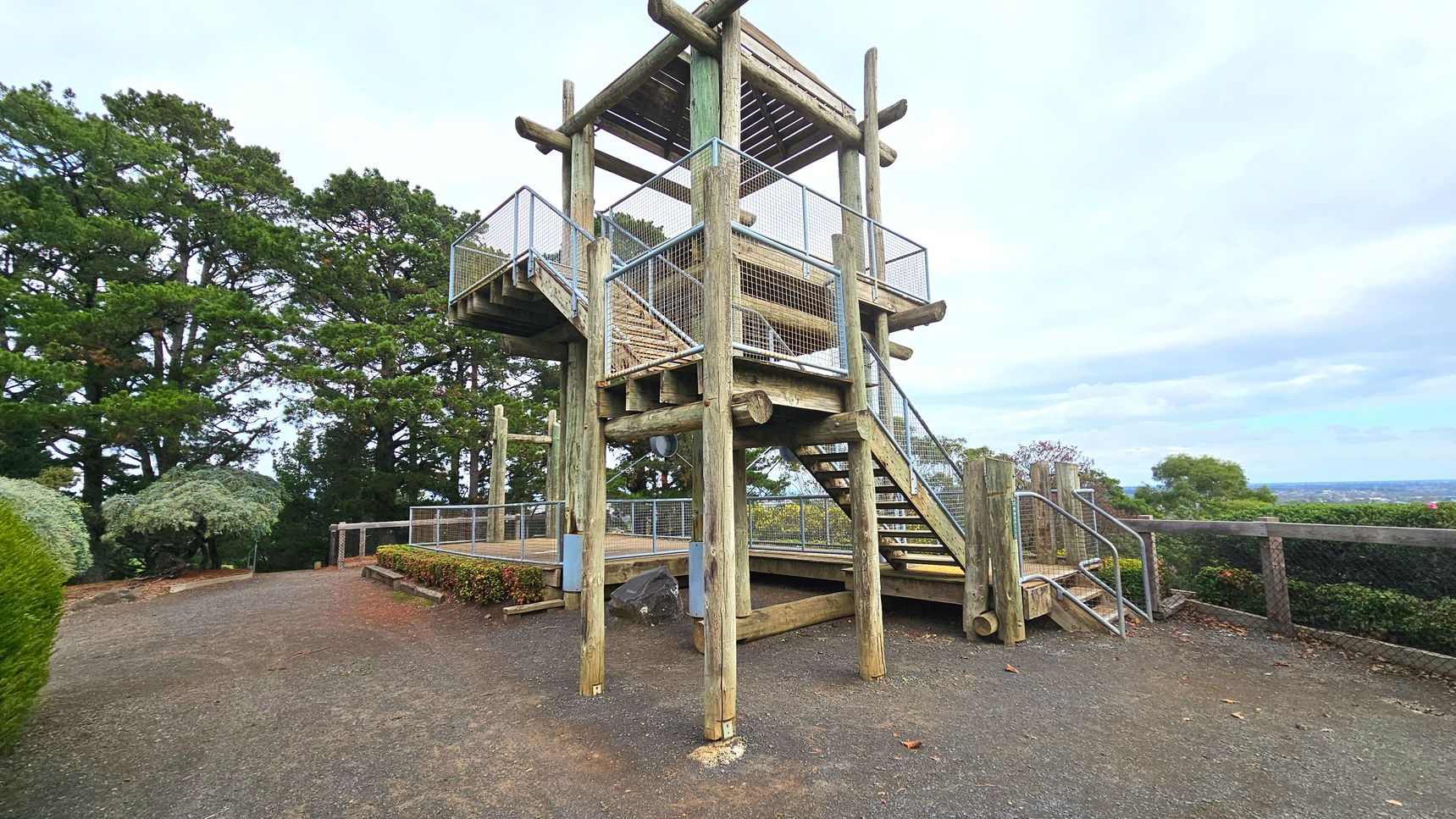 A wooden observation tower stands prominently, constructed with large timber logs and metal railings. It features a staircase leading to an upper platform, enclosed by mesh fencing. The ground is a mix of gravel and dirt, bordered by low hedges. Tall trees surround the area, providing a natural backdrop. A metal handrail is visible near the stairs, and a wooden fence lines the perimeter, offering views of the distant landscape.