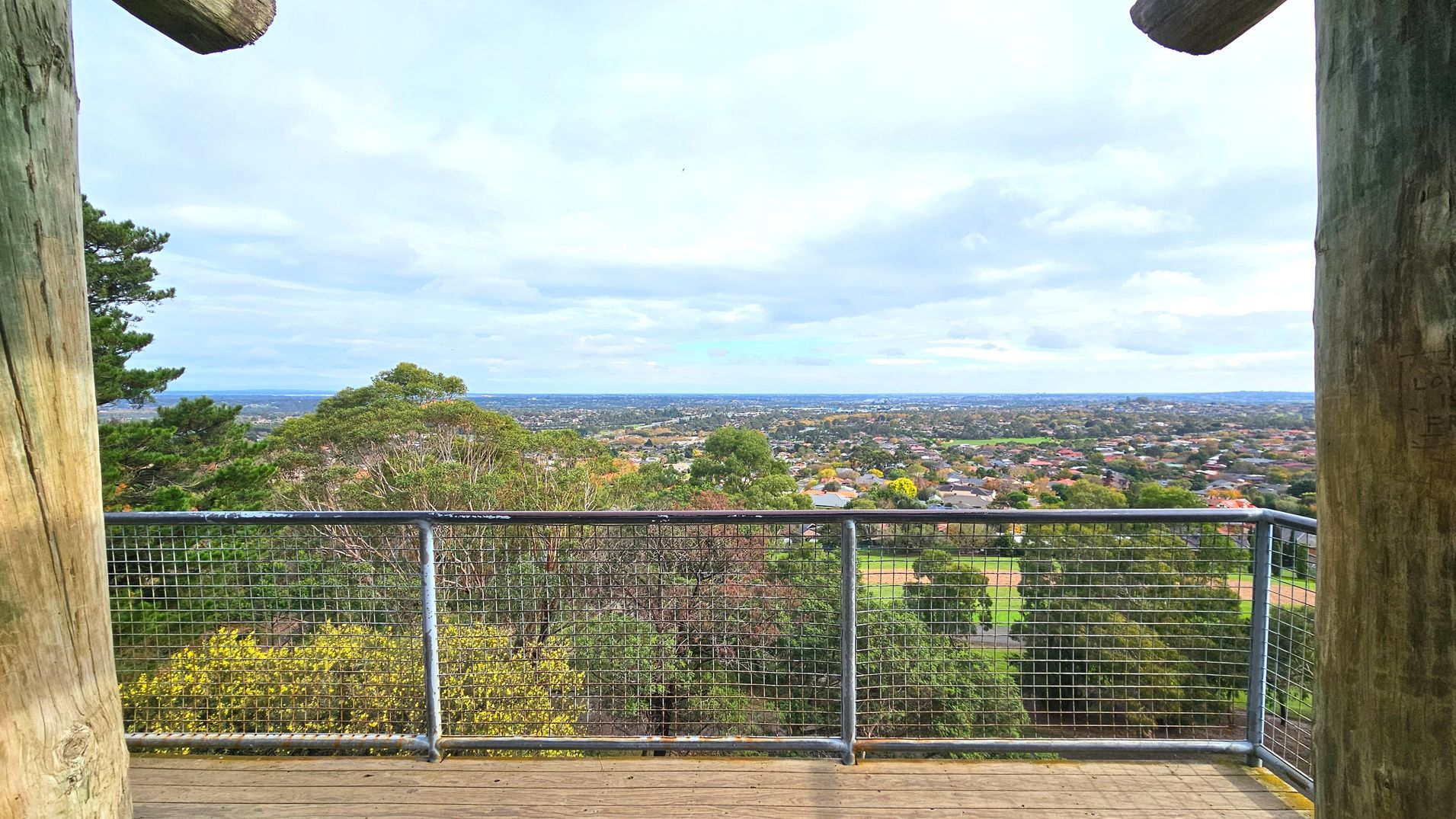 A wooden viewing platform with a metal mesh railing overlooks a sprawling suburban landscape. The platform is framed by large wooden posts on either side. Below, a variety of trees and greenery are visible, leading to a vast expanse of houses and buildings stretching to the horizon. The floor of the platform is made of wooden planks, and the railing provides a clear view of the scenery beyond.