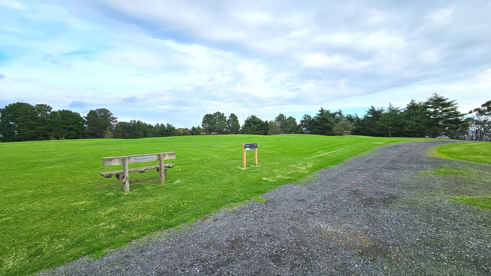 A grassy field with a wooden bench labelled 'Ball games area' on the left. A gravel path curves from the right foreground into the distance, bordered by well-maintained grass. A small wooden sign stands near the bench. In the background, a line of tall trees creates a natural boundary, giving the space an open yet enclosed feel. The grass is lush and evenly cut, suggesting a recreational area.