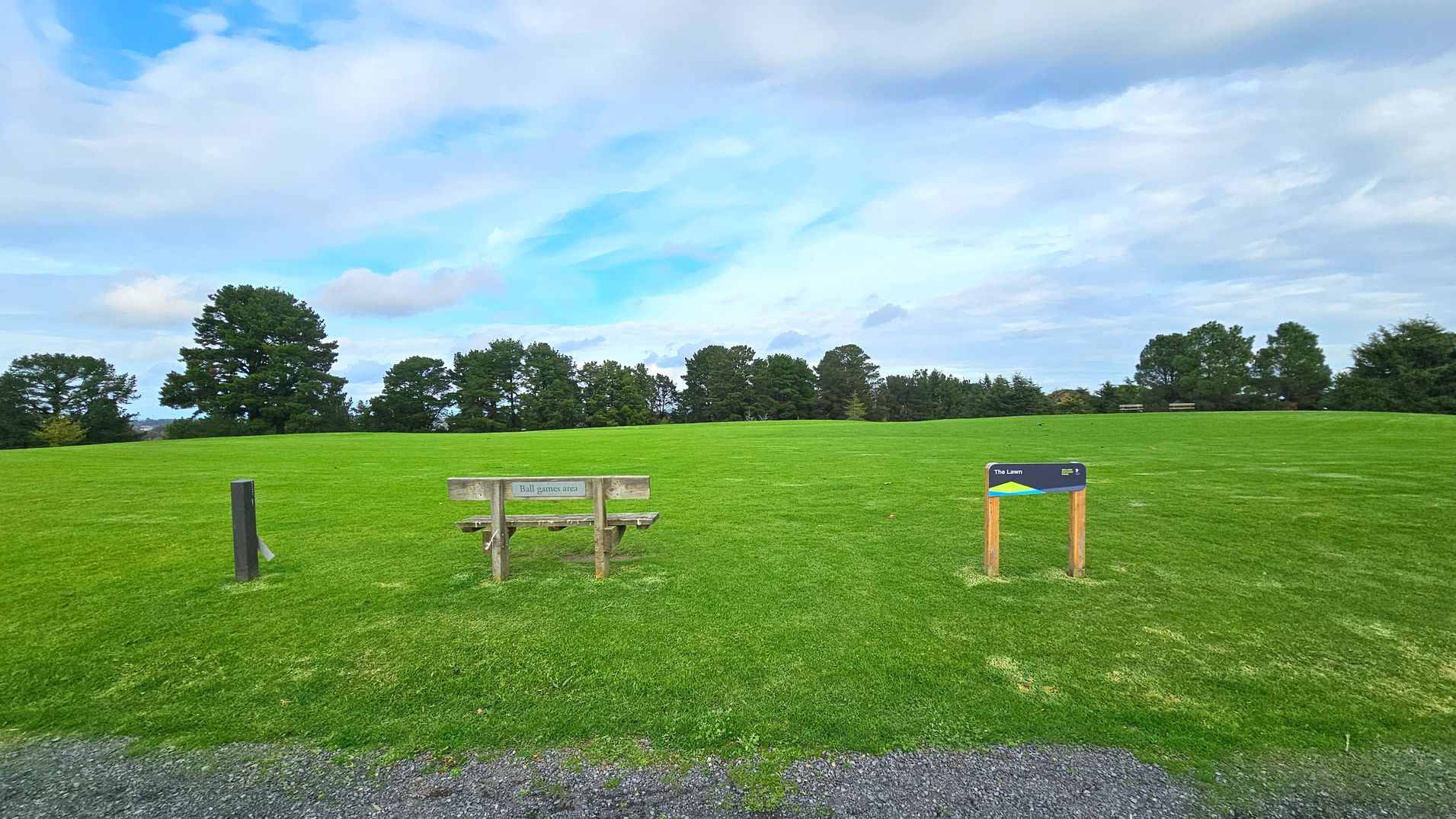 A grassy field stretches across the image, bordered by a line of trees in the background. In the foreground, a wooden bench with a sign reading 'Ball games area' is positioned to the left, while a smaller sign on wooden posts labelled 'The Lawn' stands to the right. The ground transitions from a gravel path to the lush green grass of the field, creating an open and expansive feel.