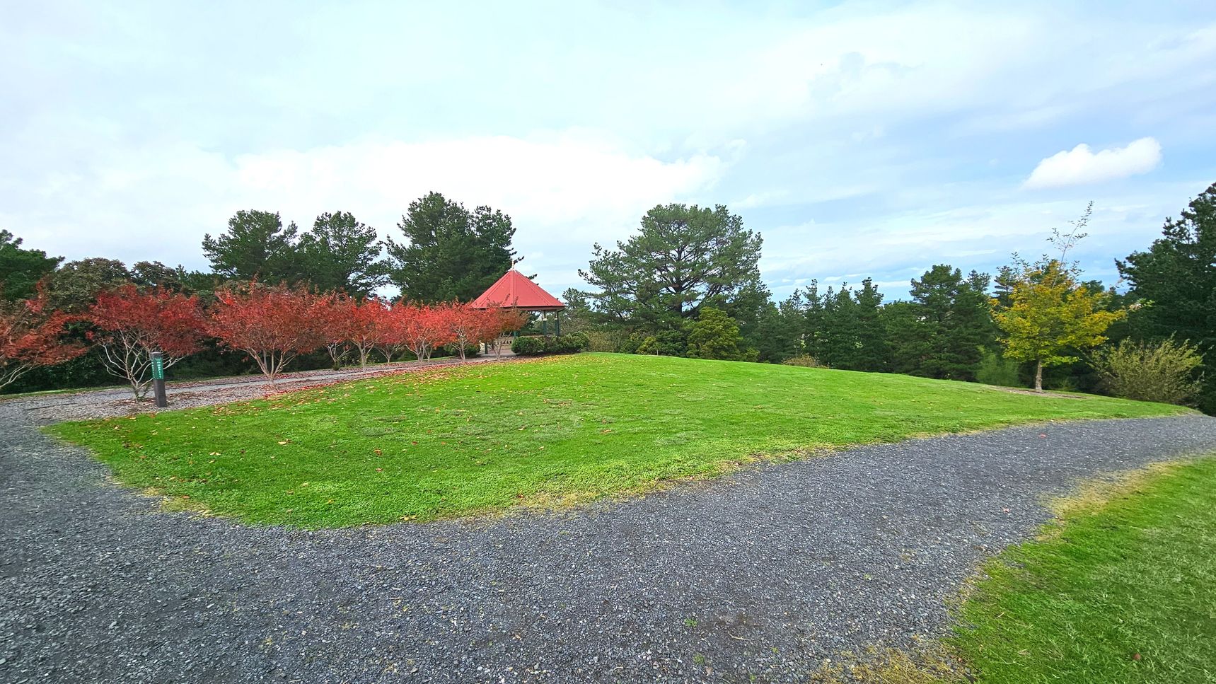 A grassy area with a gently sloping hill is bordered by a gravel path that curves around it. To the left, a row of small trees with red leaves lines the path, leading to a gazebo with a red roof partially obscured by larger green trees. The scene is surrounded by dense foliage, creating a sense of enclosure. The grass is well-maintained, and the gravel path provides a clear walking route.