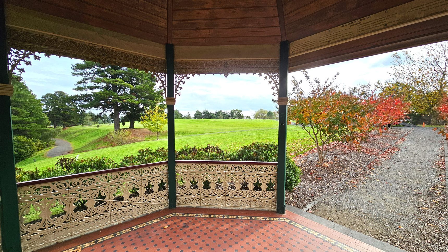 A view from inside a gazebo with ornate iron railings and a patterned tile floor. The gazebo opens onto a grassy park with a winding path and scattered trees. To the right, a gravel path lined with red-leaved bushes leads away. The gazebo's roof is wooden with decorative trim. The scene is open and expansive, with greenery and a few autumnal trees visible.