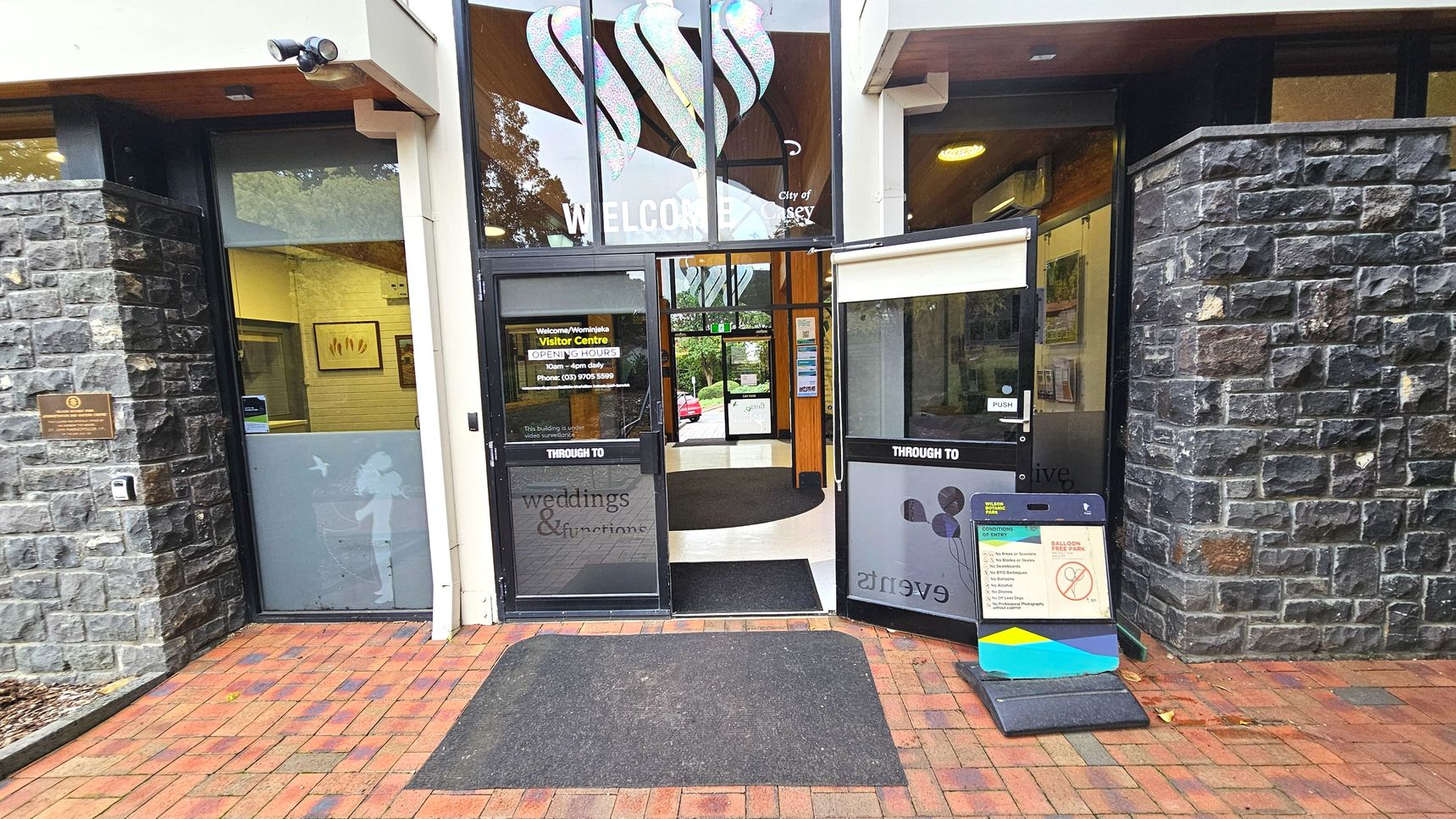 The entrance to a visitor centre features two large glass doors, one open, with signage indicating opening hours and directions to weddings and events. The facade is flanked by dark stone walls. A black mat lies on the red brick pavement leading inside. Above the doors, a decorative glass panel displays the word 'WELCOME'. A small signboard with visitor information is placed on the right side of the entrance.