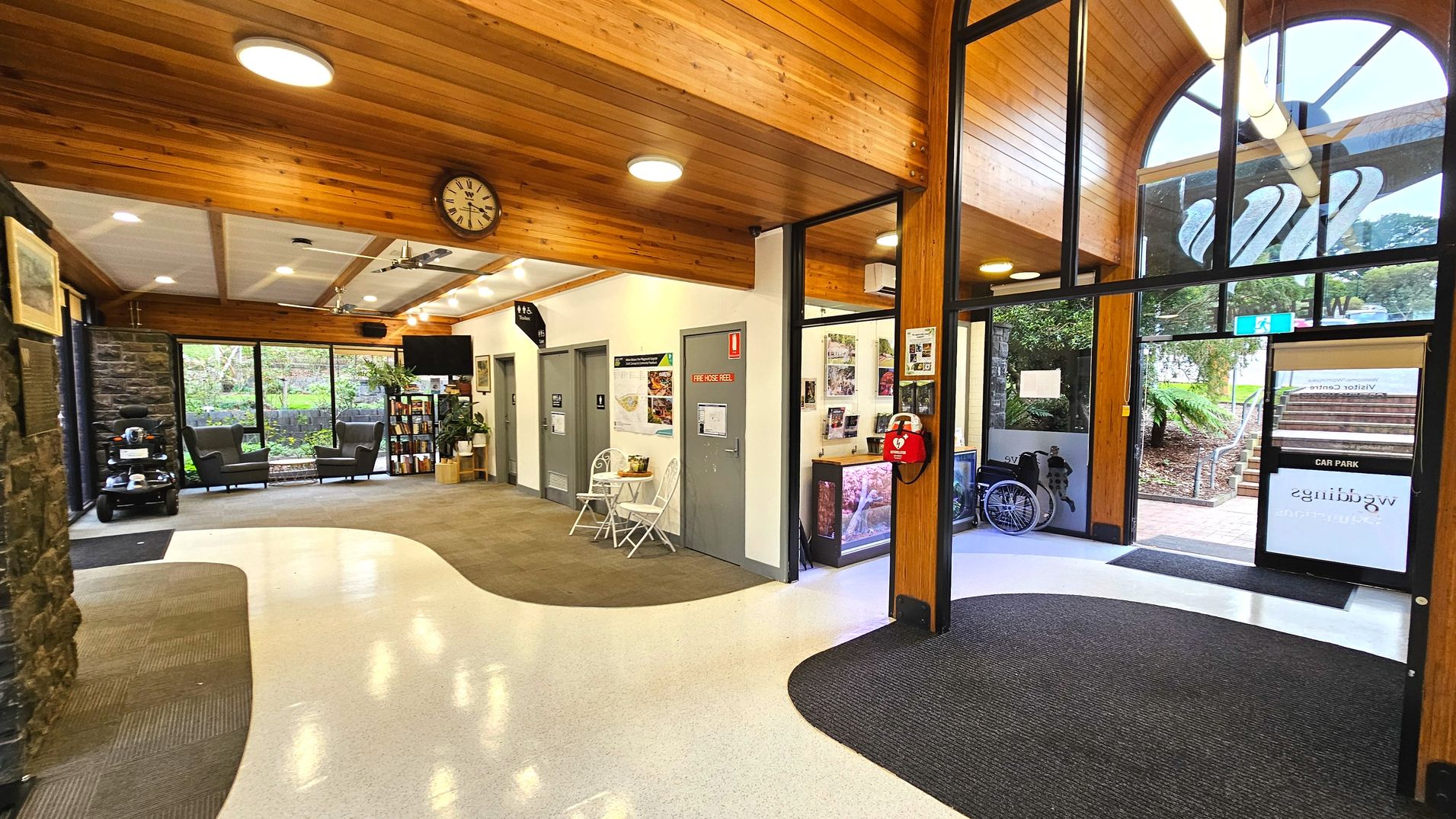 The interior of a visitor centre features a high wooden ceiling with recessed lighting and a large arched window. A clock is mounted on the wall. The floor transitions from a speckled white surface to a grey carpet. A fire hose reel is mounted on a wall near a small table with two chairs. A glass door leads outside, with a wheelchair visible nearby. A stone wall and a bookshelf with armchairs are also present.