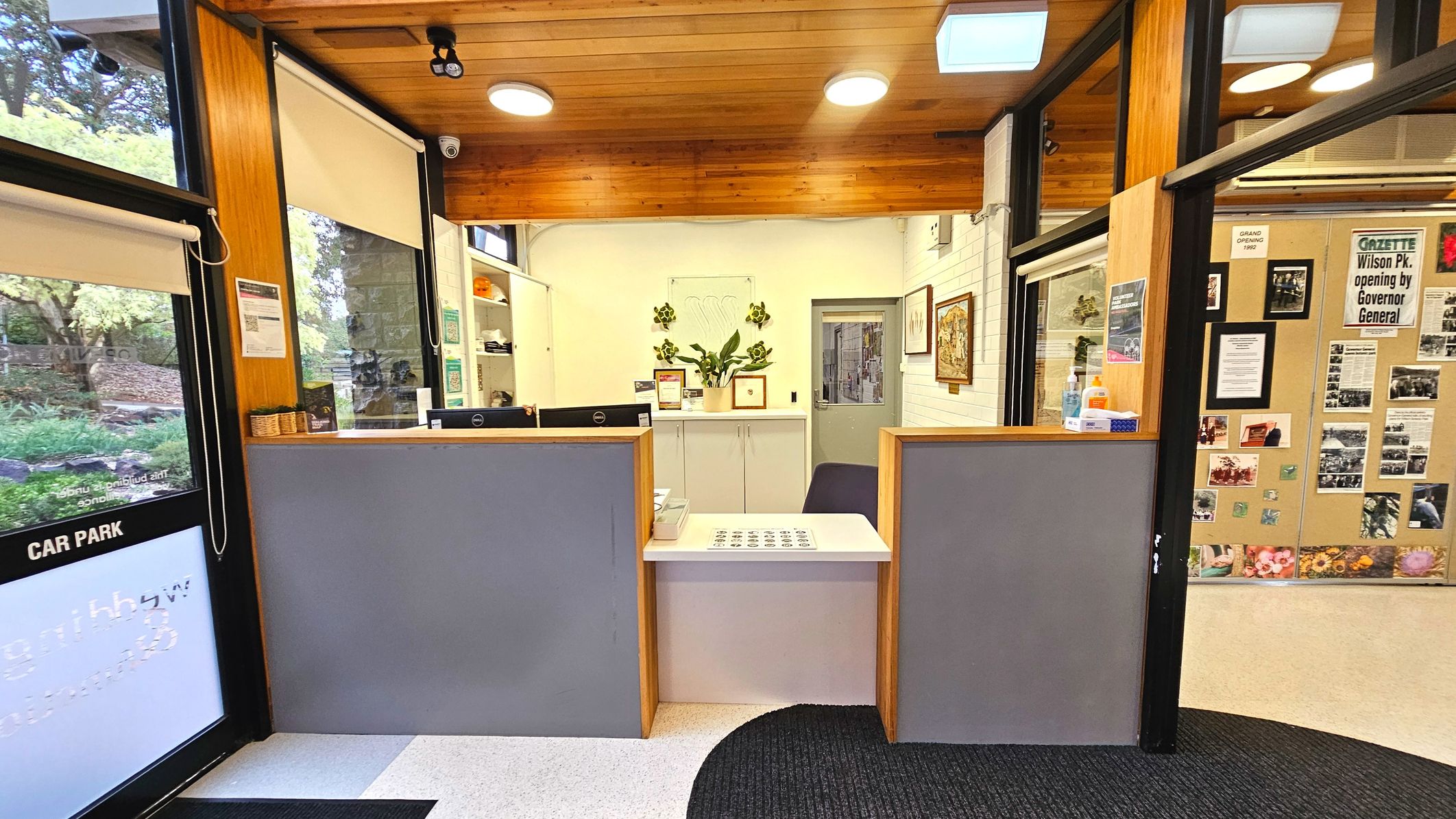 A reception area with a wooden ceiling and white walls. Two grey reception desks with wooden frames face the entrance, separated by a small white counter. Behind the desks, a white cabinet with plants and framed certificates is visible. To the right, a bulletin board displays various notices and photos. The floor transitions from a light speckled surface to a dark carpet. Large windows with roller blinds are on the left.
