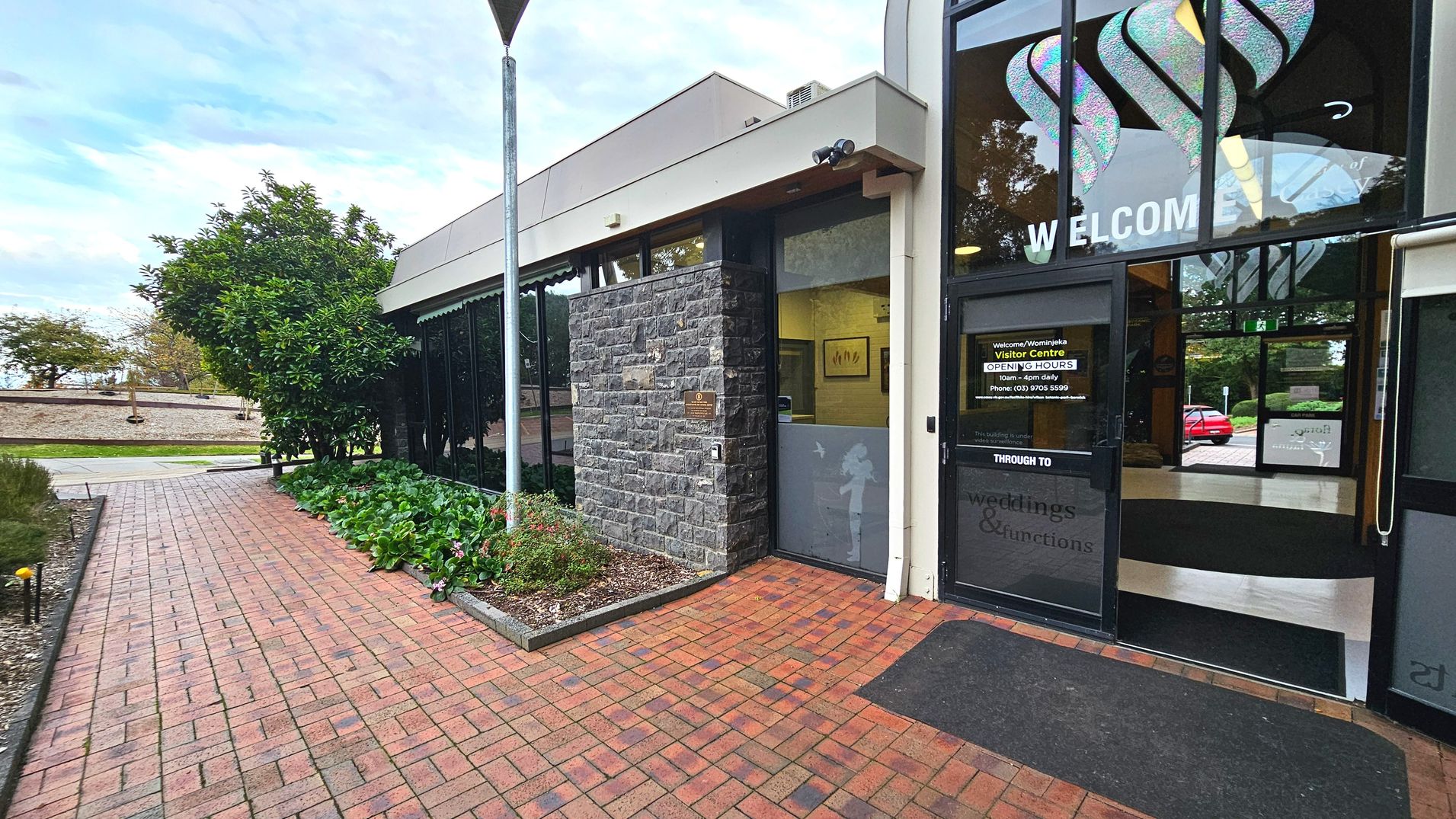 The image shows the entrance to a visitor centre with a modern design. A brick pathway leads to the glass door, which has signage indicating opening hours and directions to weddings and functions. A stone wall with a plaque is adjacent to the door. Large windows display a 'Welcome' sign. The area is landscaped with green plants and a tree, and a black mat lies at the entrance.