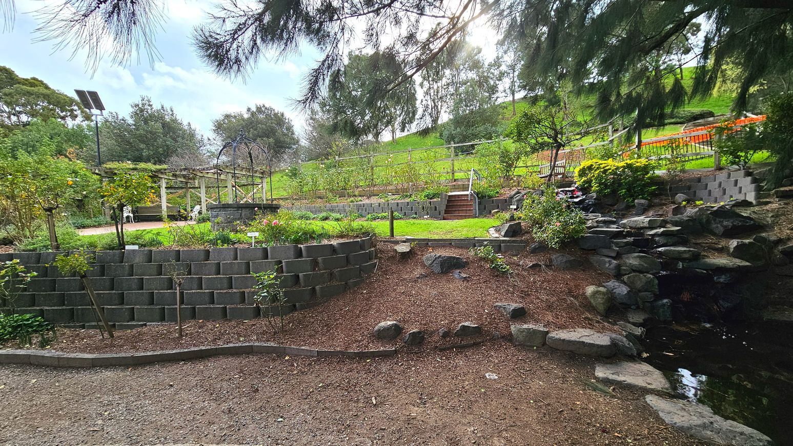 A terraced garden with multiple levels of stone retaining walls and lush greenery. In the foreground, a dirt path leads to a small pond bordered by rocks. Mid-ground features a grassy area with a wooden pergola and white chairs. Steps with a handrail ascend to higher terraces filled with plants. A solar panel on a pole is visible to the left. Trees and shrubs surround the area, creating a natural enclosure.