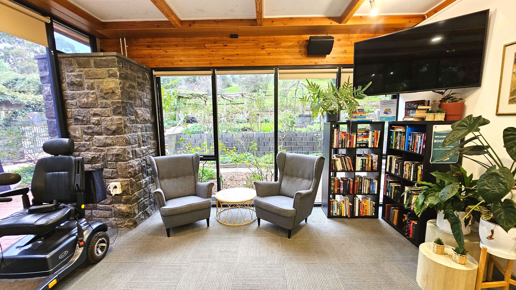 A cosy reading nook with two grey armchairs facing each other, separated by a small round wicker table. Behind them, large floor-to-ceiling windows offer a view of a lush garden. To the right, a black bookshelf filled with books stands beneath a large wall-mounted TV. A mobility scooter is parked to the left near a stone wall. The floor is carpeted with a subtle grid pattern, and several potted plants add greenery to the space.