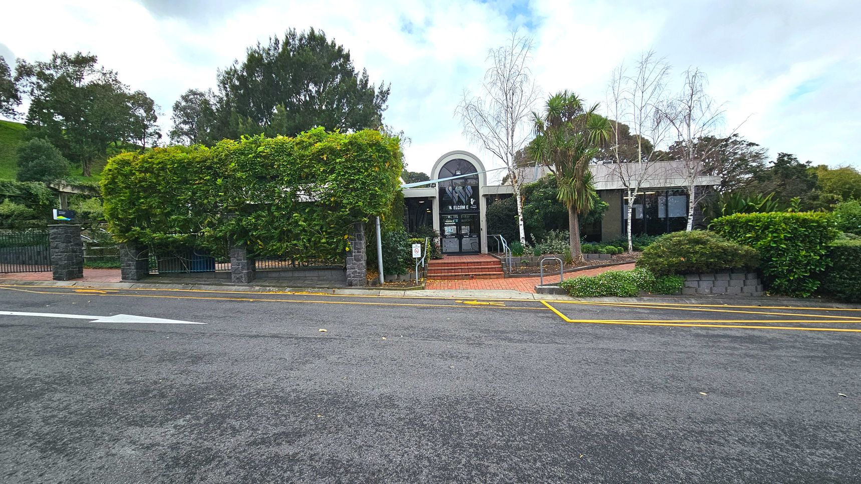A building with a large arched entrance is partially obscured by lush greenery and trees. The entrance features glass doors and a 'Welcome' sign. Brick steps lead up to the entrance, flanked by metal railings. The foreground shows a paved road with yellow parking lines and a directional arrow. Stone pillars and a metal gate are visible to the left, with dense foliage surrounding the area.