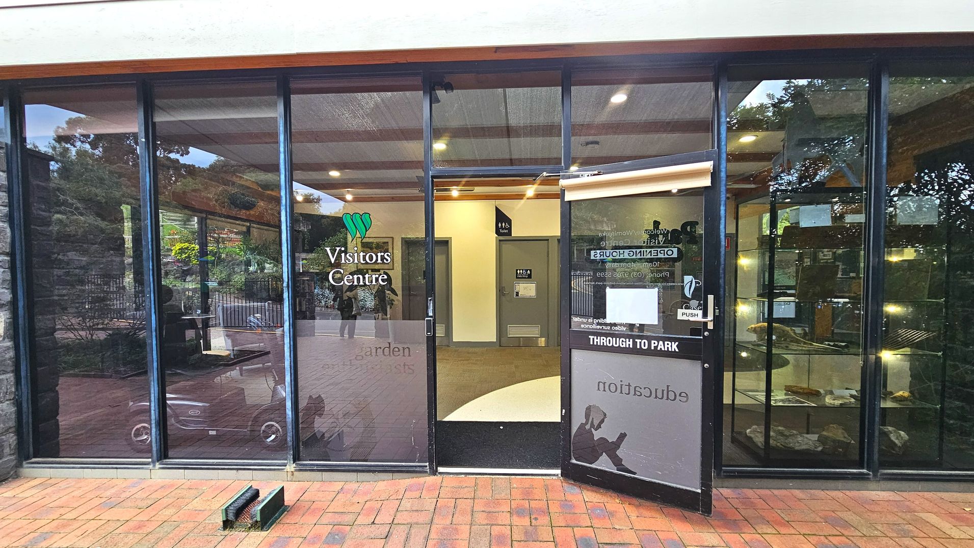 The entrance to a Visitors Centre features large glass panels with a door slightly ajar. The glass displays the centre's name and opening hours. Inside, a beige carpet leads to a door marked 'Toilet'. The exterior has a brick pavement, and a shoe brush is placed near the entrance. The interior is well-lit with ceiling lights, and a display case with various items is visible through the glass.