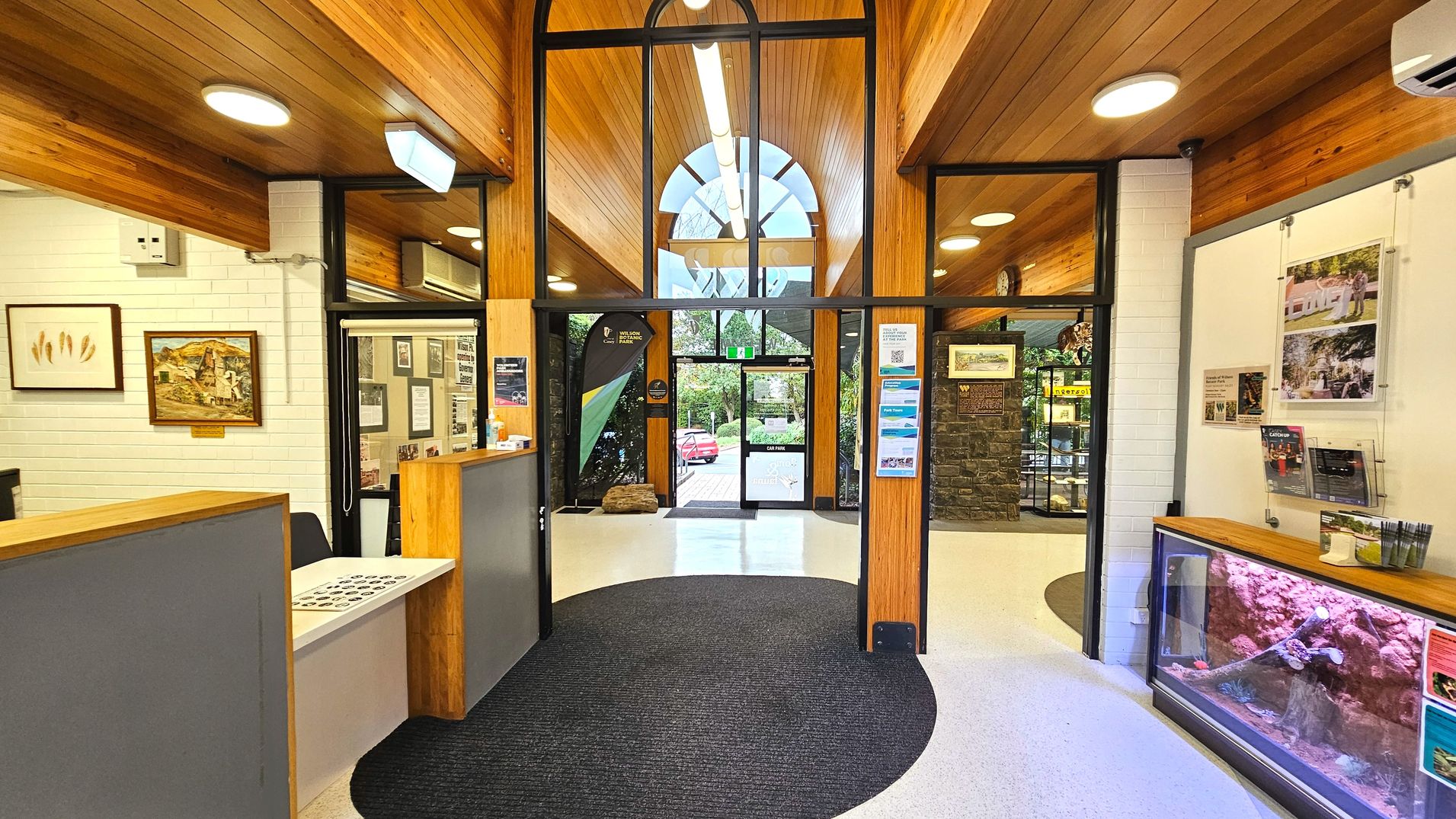 The interior of a visitor centre features a high wooden ceiling with recessed lighting and a large arched window above the entrance. The space is enclosed by white brick walls and glass panels. A reception desk with brochures is on the left, and a display case with rocks and informational posters is on the right. The floor transitions from white tiles to a dark circular carpet near the entrance, which leads to a glass door with a green exit sign.