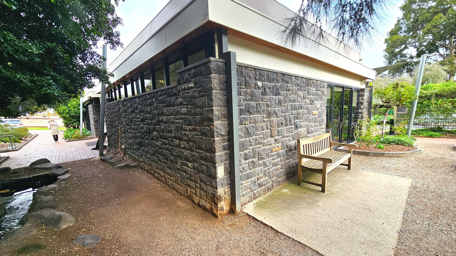 A stone building with a flat roof and narrow windows runs diagonally across the image. A wooden bench sits on a concrete slab near the building's corner. A paved path leads away from the building, bordered by greenery and a small pond with rocks. A person in a coat walks along the path. The area is surrounded by trees and shrubs, creating a semi-enclosed garden-like setting.
