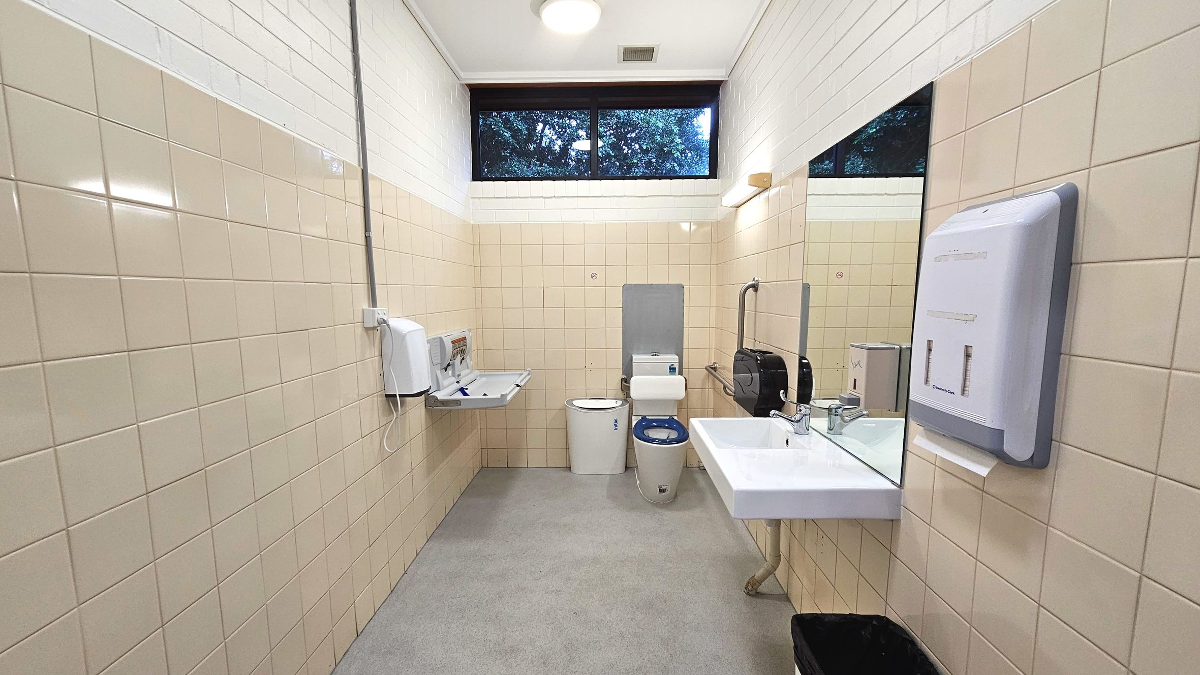 A public restroom with beige tiled walls and a grey tiled floor. A white toilet with a blue seat is positioned against the back wall, flanked by a baby changing station and a sanitary bin. A white sink with a mirror above it is on the right wall, accompanied by a soap dispenser and a hand dryer. A black waste bin is on the floor. A high window with a view of trees provides natural light.