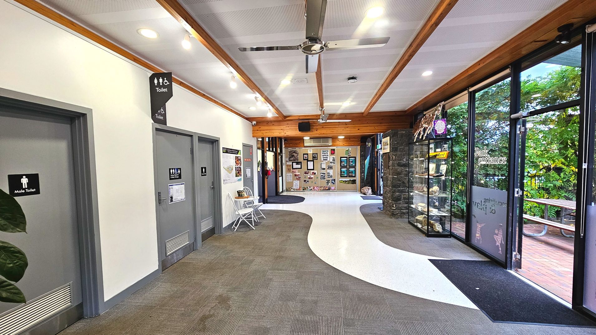 A hallway with a mix of carpet and smooth white flooring, leading to a glass entrance with 'Park entrance' signage. On the left, grey doors marked 'Male Toilet' and 'Toilet' are visible. A small white table and chair set is placed nearby. The right side features a stone wall with display cases containing various items. The ceiling has wooden beams and metal fans, and outside, a picnic table is visible through the glass.