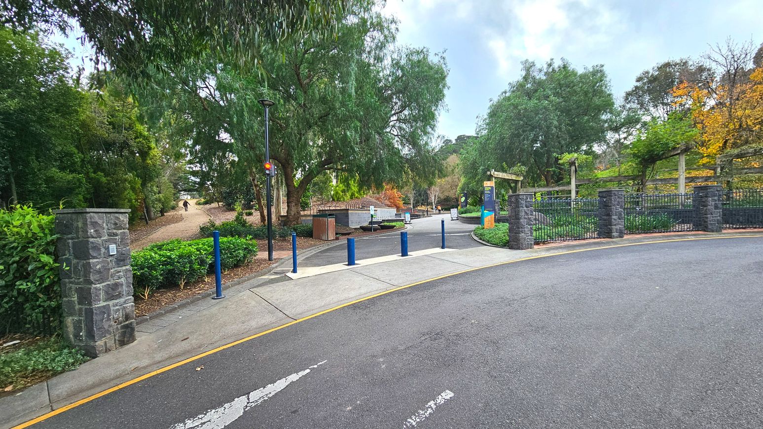 A paved road curves gently, bordered by a low stone wall and metal fence on the right, leading into a park area. Blue bollards line the entrance, with a pedestrian path veering left into a wooded area. A tall lamp post stands near the path, and a large tree provides shade. Signage is visible near the entrance, and the area is surrounded by lush greenery and trees, creating a semi-enclosed feel.