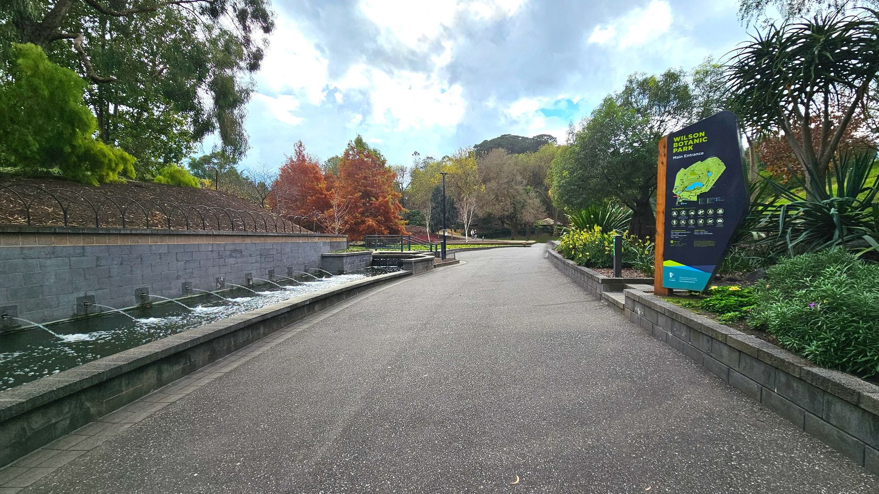 A paved pathway curves gently through a landscaped park area. On the left, a low stone wall with water features spouts streams into a narrow pool. To the right, a large informational sign for Wilson Botanic Park stands amidst lush greenery and flowering plants. Trees with autumnal foliage line the path, and a lamppost is visible further along the path, which is bordered by low stone walls.