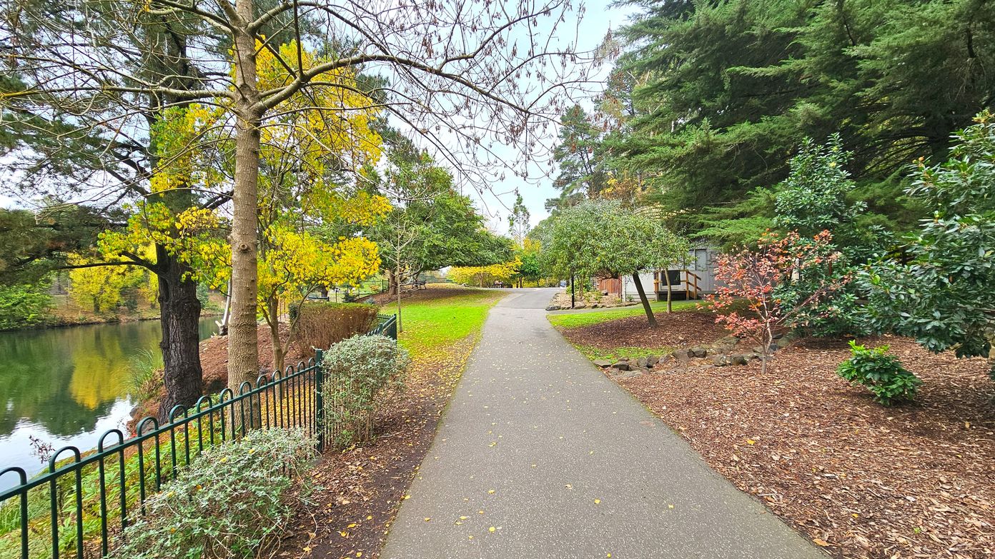 A paved path curves gently through a park, bordered by a black metal fence on the left, separating it from a calm body of water. Trees with autumn foliage line both sides, with a mix of yellow, green, and red leaves. The ground is covered with mulch and grass, and a small building is partially visible in the background. The area feels open with a clear view of the path and surrounding greenery.