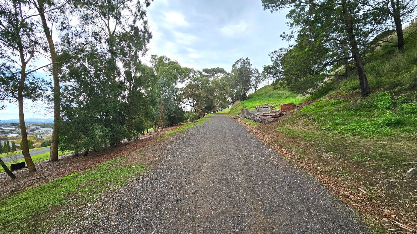 A gravel path stretches into the distance, flanked by tall trees on both sides. To the right, a grassy slope rises, dotted with rocks and a small brick structure. On the left, the land slopes downwards, revealing a view of a distant town. The path is bordered by patches of green grass and brown earth, creating a natural, open environment with no visible barriers.