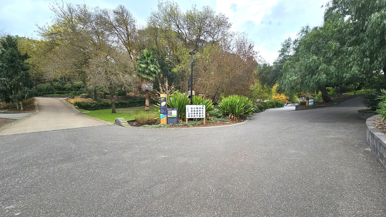 A paved pathway forks into two directions surrounded by lush greenery and trees. In the centre, a tall black lamppost stands beside a wooden signboard and a white information board with circular symbols. The left path is slightly elevated with a gentle incline, while the right path curves gently. The area is bordered by low stone walls and dense foliage, creating a sense of enclosure.
