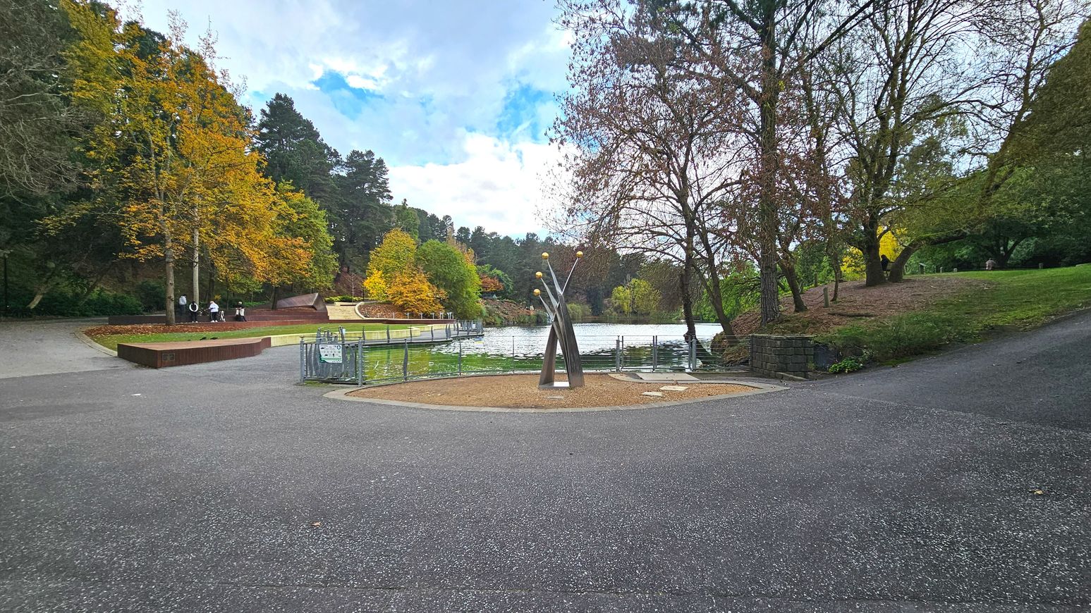 A park scene with a central metal sculpture featuring three tall, intersecting poles topped with spherical elements. Surrounding the sculpture is a circular gravel area bordered by a low metal fence. To the left, a paved path leads past autumnal trees and a wooden bench. A lake with a railing runs along the background, flanked by lush greenery and trees. The area feels open with gentle slopes and varied pathways.