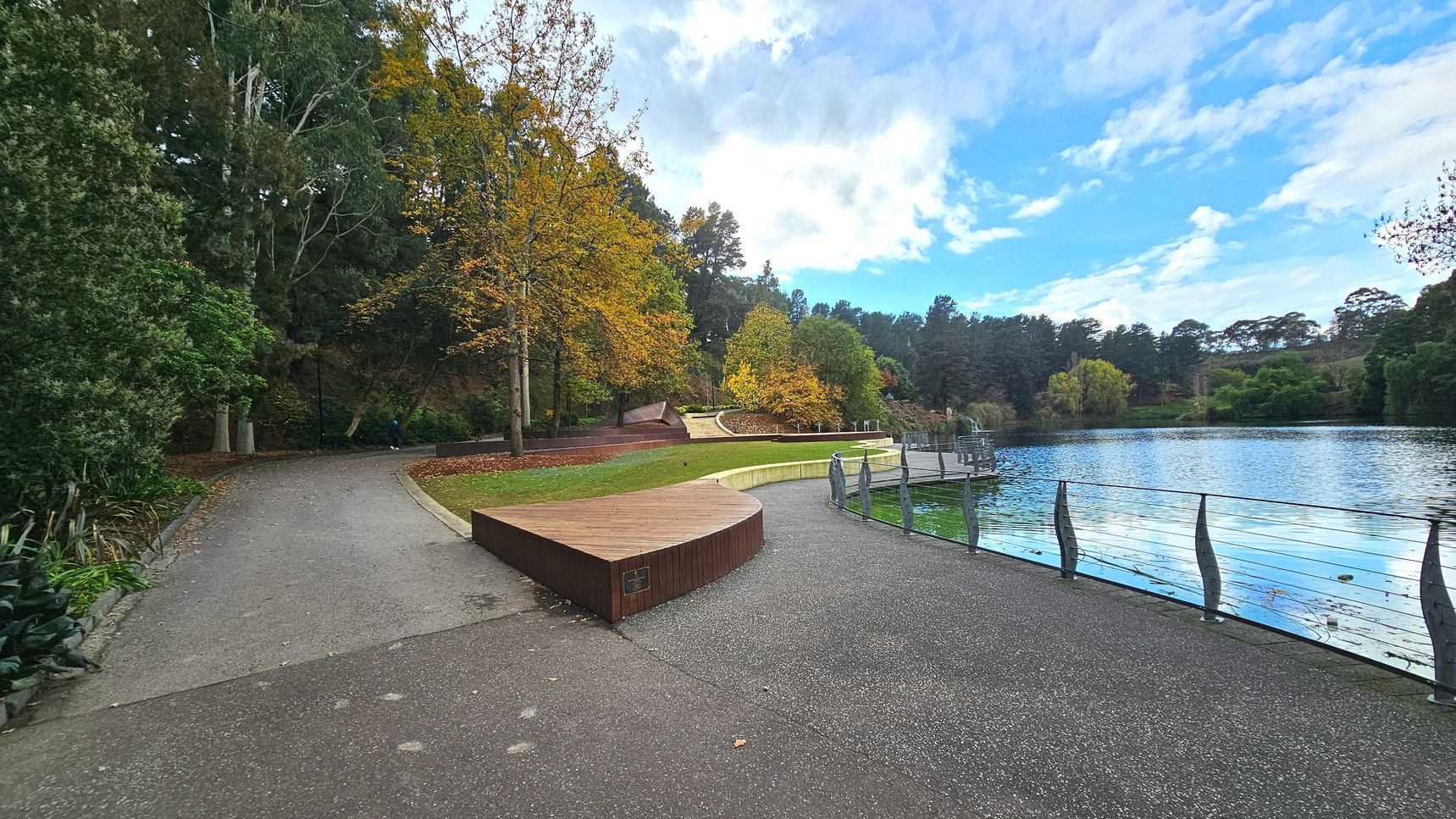 A serene park scene features a curved wooden bench on a paved path beside a tranquil lake. The path curves gently to the left, bordered by lush greenery and tall trees with autumnal leaves. A metal railing with artistic vertical supports lines the lake's edge. The area feels open, with a grassy section leading to more trees and a small dock extending into the water.