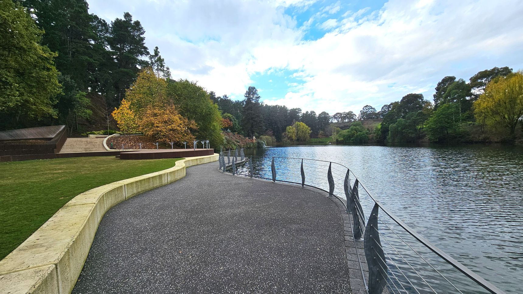 A curved pathway with a textured surface runs alongside a large body of water, bordered by a metal railing with decorative elements. To the left, a grassy area is edged by a low stone wall, leading up to a series of steps and landscaped areas with trees in autumn colours. The scene is framed by dense trees in the background, creating a natural enclosure around the water and path.