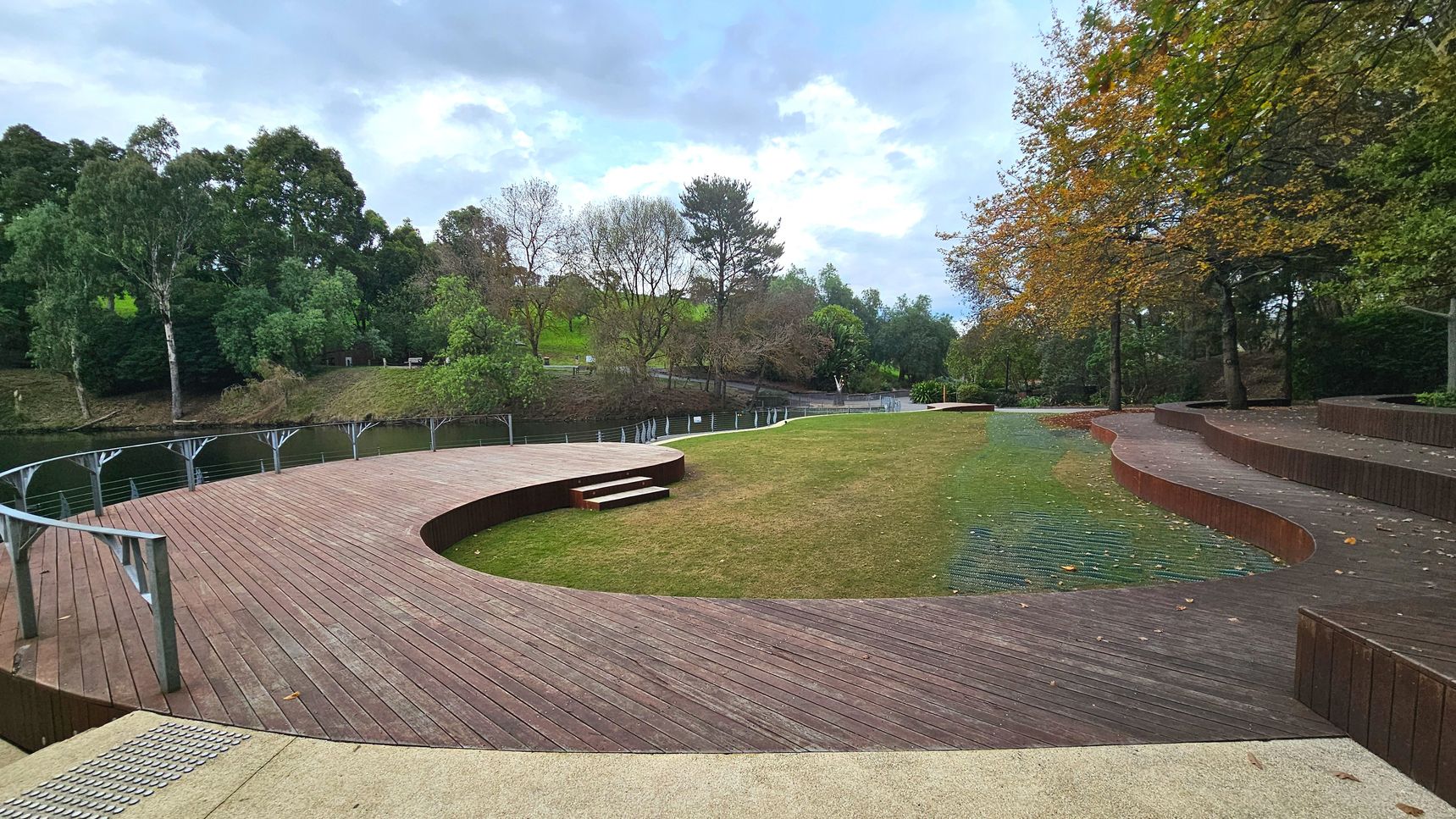 A curved wooden deck with metal railings overlooks a grassy area, bordered by a series of tiered wooden seating steps. The deck features a small set of steps leading down to the grass. Trees with green and autumnal leaves line the right side, while a body of water is visible on the left, separated by a metal fence. The space feels open, with a mix of natural and constructed elements.