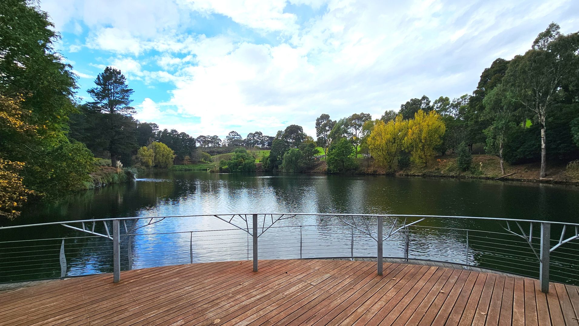 A wooden deck with metal railings overlooks a tranquil lake surrounded by lush greenery. The deck features evenly spaced vertical metal posts with decorative elements. The lake is bordered by a variety of trees, including some with yellowing leaves, suggesting a seasonal change. The scene is serene, with the water reflecting the surrounding foliage. The area appears open and inviting for visitors.