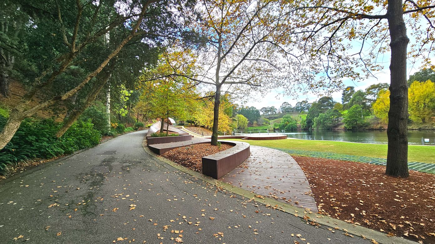 A winding paved path curves through a park, bordered by lush greenery and trees with autumn leaves. To the right, a series of curved wooden benches are integrated into the landscape, surrounded by fallen leaves. Beyond the benches, a grassy area leads to a calm river, with trees lining the opposite bank. The scene is serene, with a mix of natural and landscaped elements.
