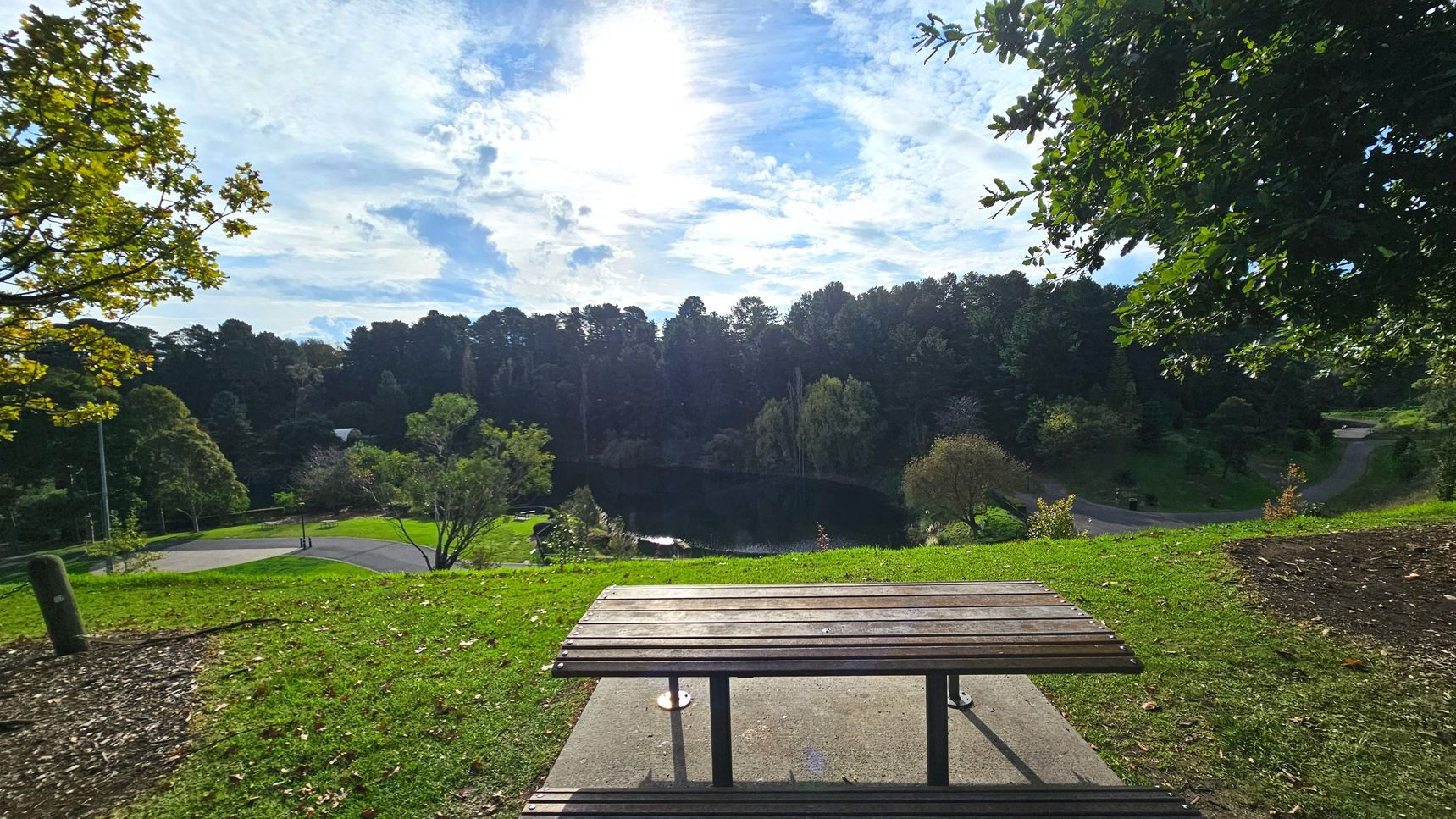 A wooden picnic table is positioned on a grassy area overlooking a serene park landscape. In the foreground, the table rests on a concrete slab surrounded by grass. Beyond, a gently sloping hill leads down to a small pond bordered by dense trees. A paved path curves through the park, with scattered trees and shrubs adding to the natural setting. The area feels open and inviting, with lush greenery and a tranquil atmosphere.