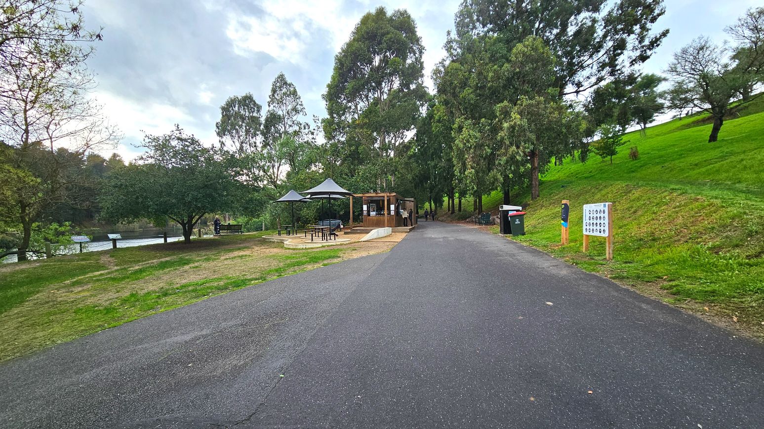 A paved path leads to a small wooden kiosk surrounded by tall trees and greenery. To the left, a seating area with picnic tables and umbrellas is visible, adjacent to a body of water. On the right, a grassy slope rises with a couple of signs and a bin near the path. The area feels open with a mix of natural and man-made elements, providing a serene setting for visitors.