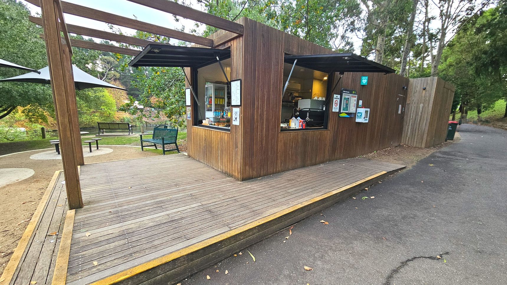 A small wooden kiosk with open serving windows is situated on a wooden deck. The kiosk has a simple, modern design with vertical wooden panels and a flat roof. Nearby, there are green metal benches and round concrete tables on a dirt surface. A path made of asphalt runs alongside the kiosk, bordered by trees and greenery. The area feels open with a mix of natural and constructed elements.
