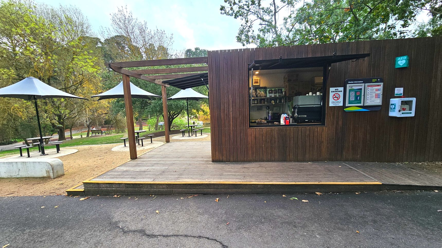 A small wooden kiosk with a serving window is situated on a wooden deck. The kiosk has a coffee machine visible inside and various signs, including a no smoking sign and an AED unit on the wall. Adjacent to the kiosk, there are several round tables with black metal umbrellas on a sandy surface. The area is surrounded by trees and greenery, creating a park-like setting. The ground transitions from asphalt to wood decking.