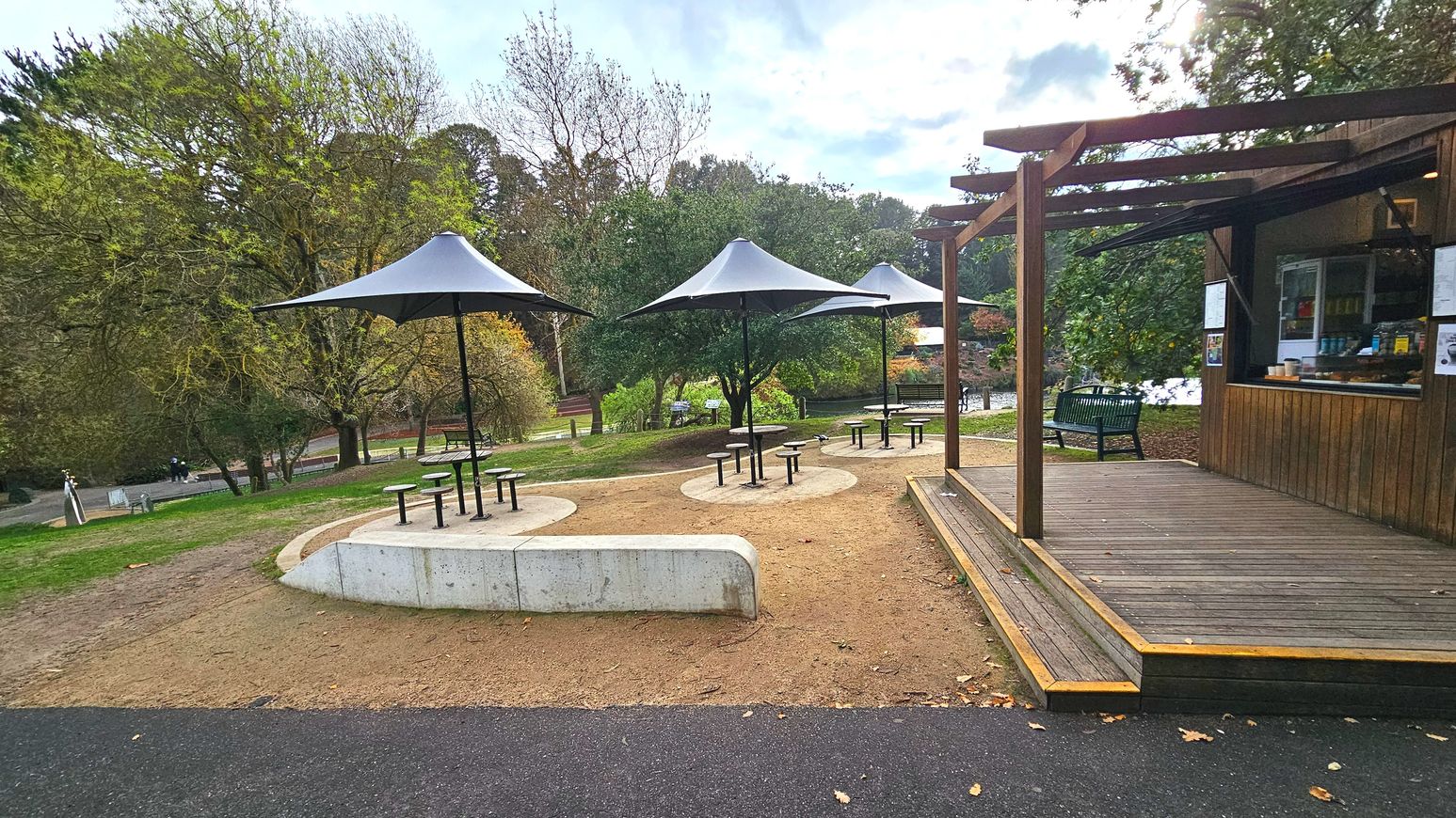 A small outdoor seating area with three round tables, each shaded by a large, conical umbrella. The tables are surrounded by stools and set on circular concrete pads. Adjacent is a wooden kiosk with a serving window, sheltered by a pergola. The ground transitions from a paved path to sandy soil. Trees with autumn foliage surround the area, providing a natural backdrop.