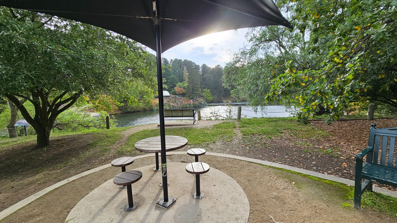 A park scene with a round wooden picnic table and four stools under a large black umbrella. The table is set on a circular concrete pad surrounded by gravel. To the right, a green wooden bench is partially visible. In the background, a serene pond is bordered by lush trees, with a wooden bench facing the water. The area is open with a mix of grass and dirt paths leading to the pond.