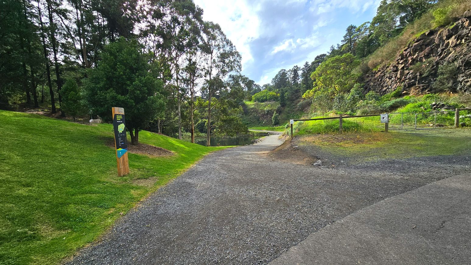 A gravel path curves gently through a lush, green parkland, flanked by well-maintained grass and dense trees. A wooden signpost with a map stands on the left, while a wooden fence with a small gate is visible on the right. The path leads towards a small pond surrounded by rocky cliffs and more greenery. The area feels open with natural boundaries formed by trees and rocks.