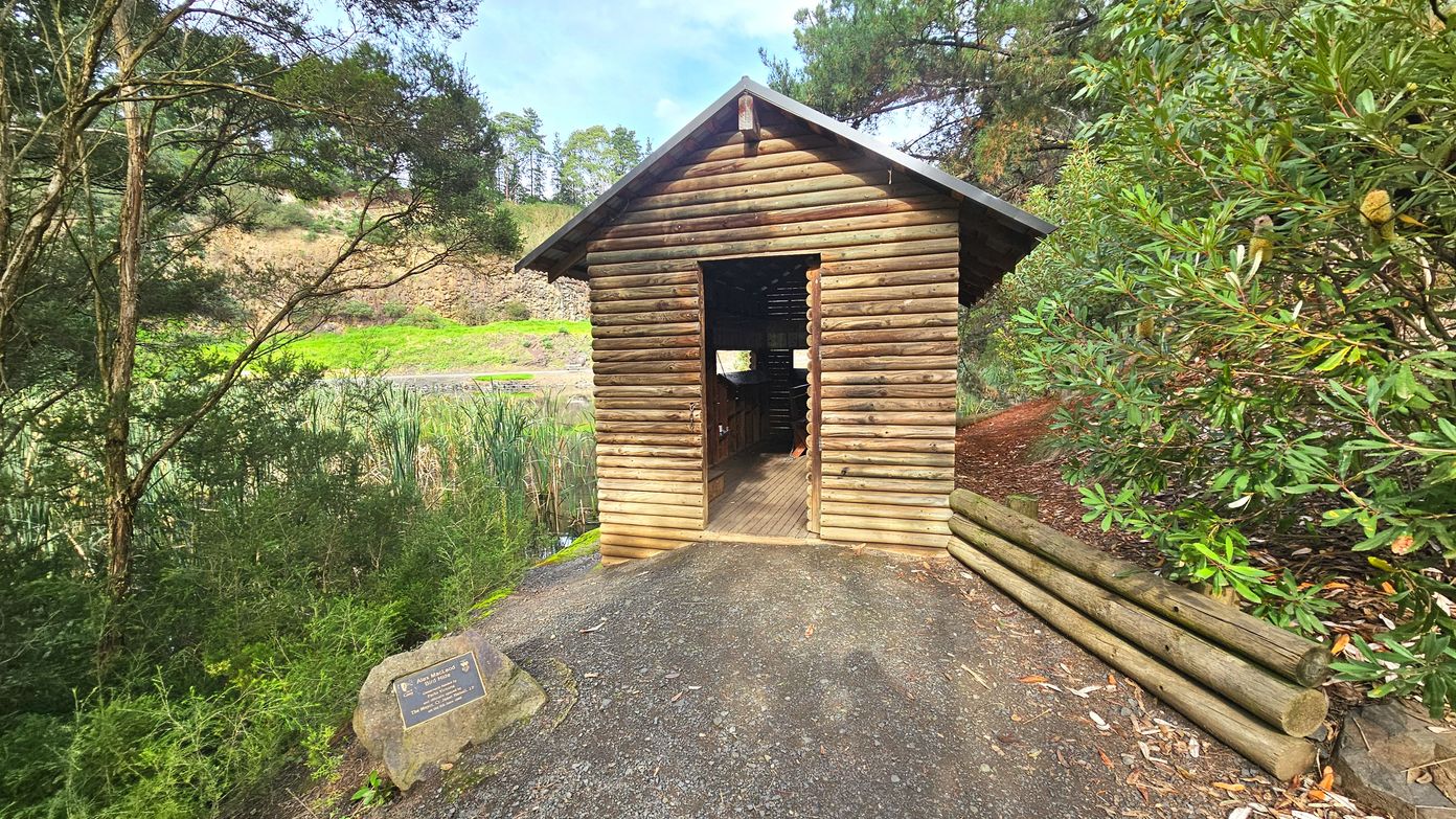 A small wooden hut with a pitched roof stands in a natural setting, surrounded by dense greenery and trees. The hut is constructed from horizontal logs and has an open doorway leading to a wooden floor inside. A stone with a plaque is positioned on the ground to the left of the hut. The area is enclosed by lush vegetation, with a gravel path leading up to the entrance.