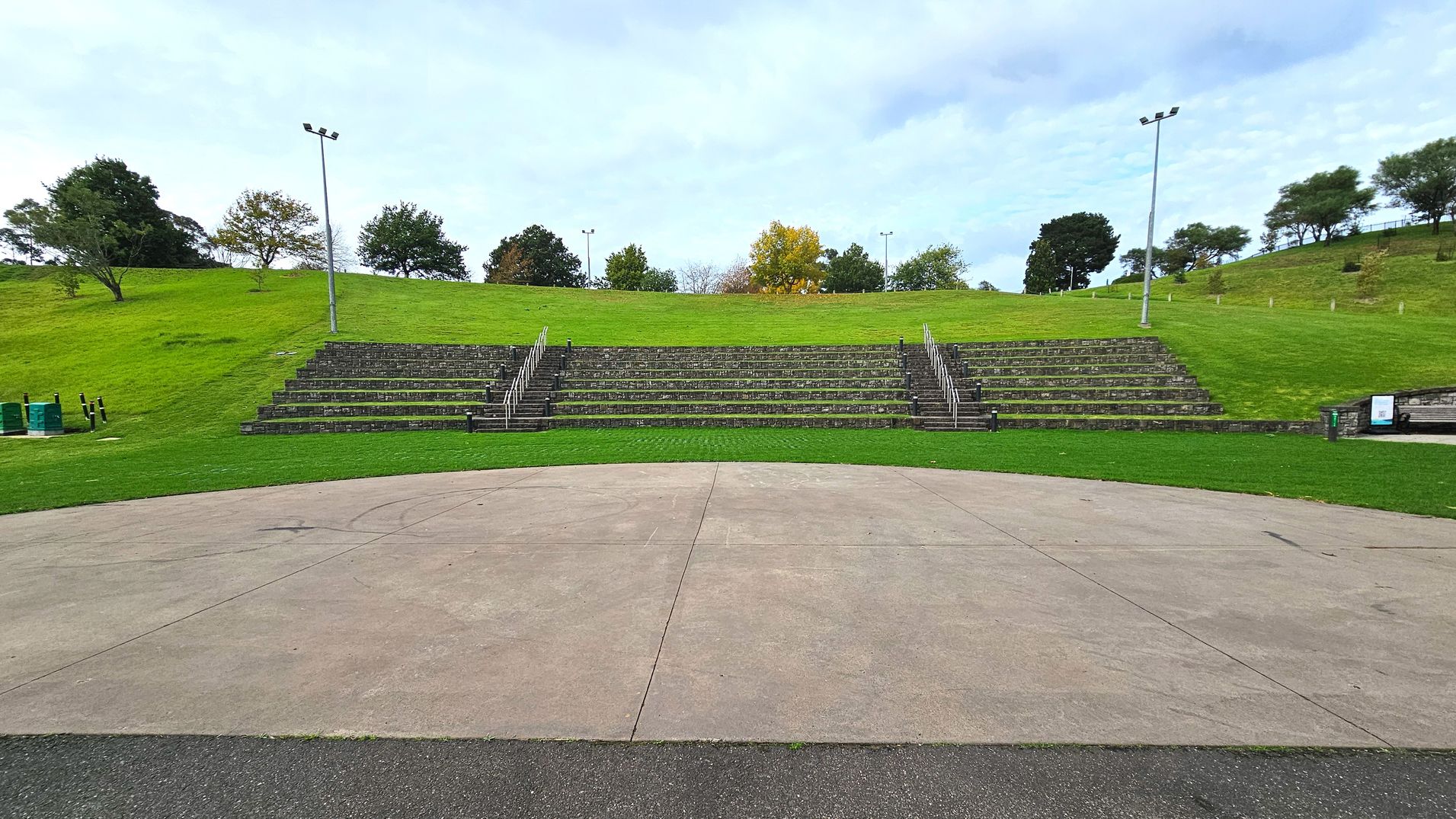 An outdoor amphitheatre with tiered stone seating set into a grassy hillside. The seating is arranged in a semi-circle with metal railings dividing sections. Two tall floodlights flank the seating area. In front, a large, flat concrete stage is bordered by a strip of asphalt. Trees are scattered across the hilltop, providing a natural backdrop. A signboard is visible to the right of the seating.